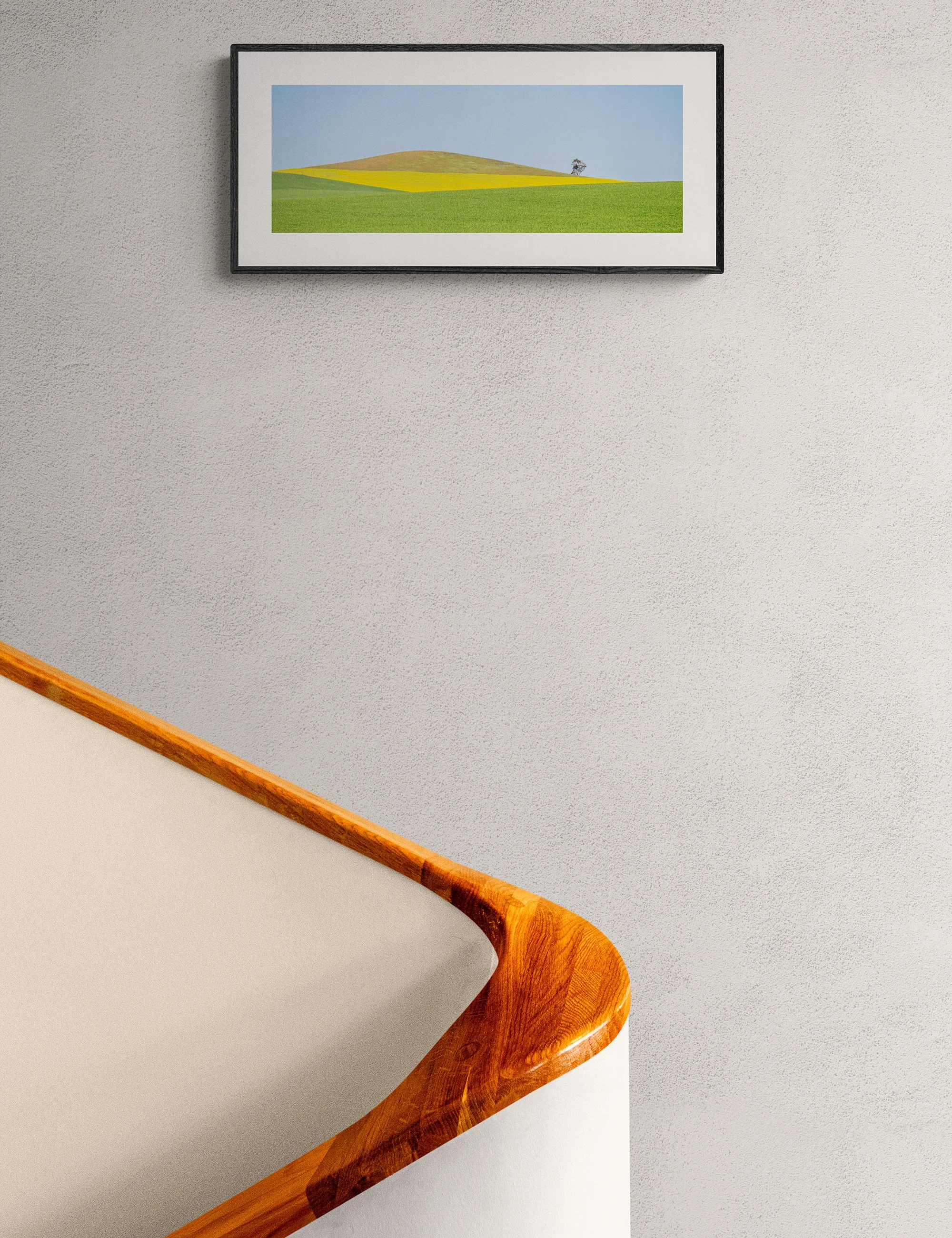 “Lone Tree Sentinel” framed minimalist landscape print by Ian J. Muirhead Arts above a curved timber staircase in a minimalist interior. Rolling green and golden fields surround a solitary tree.