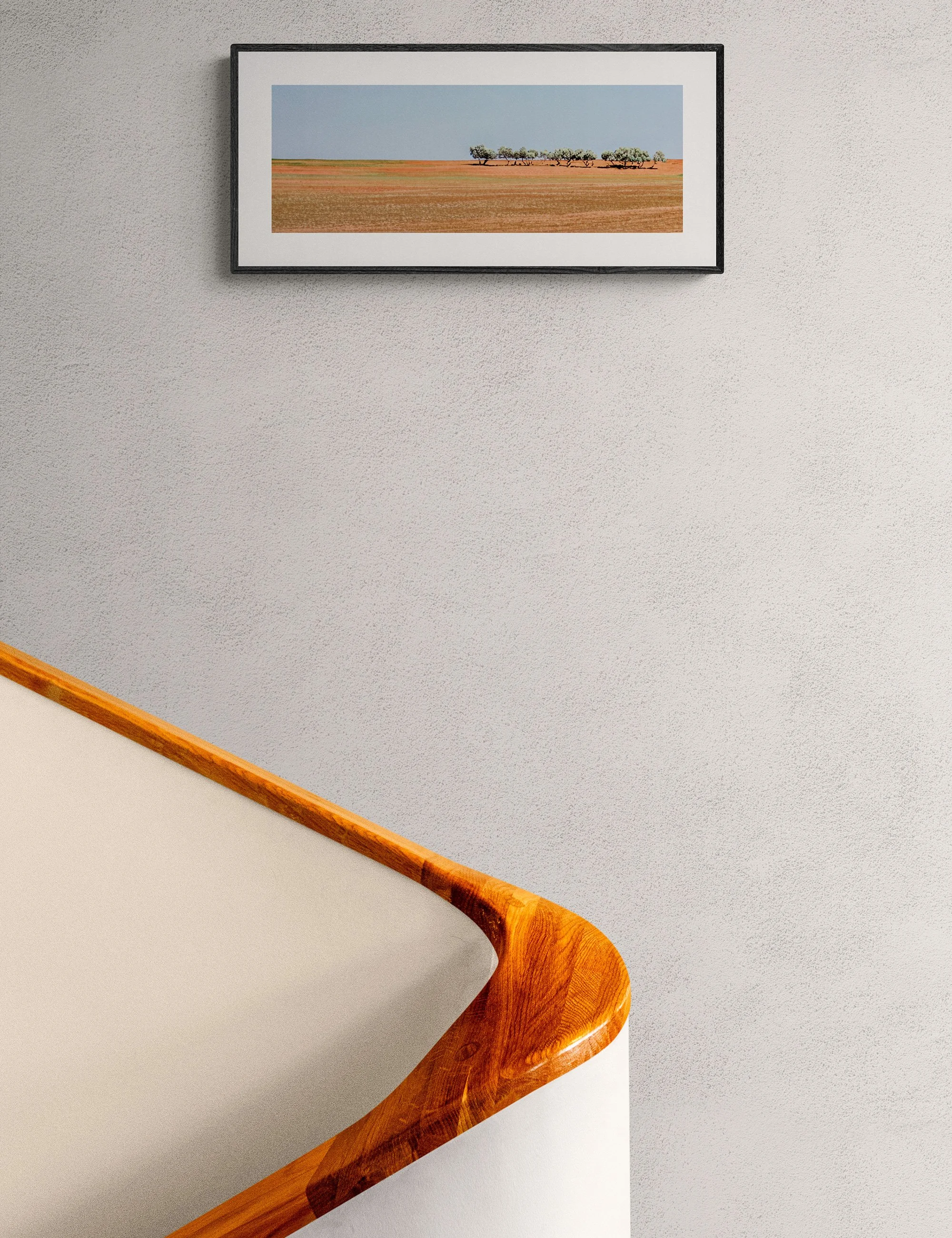 “After the Wheat” framed landscape print by Ian J. Muirhead Arts above a curved timber staircase in a minimalist interior.