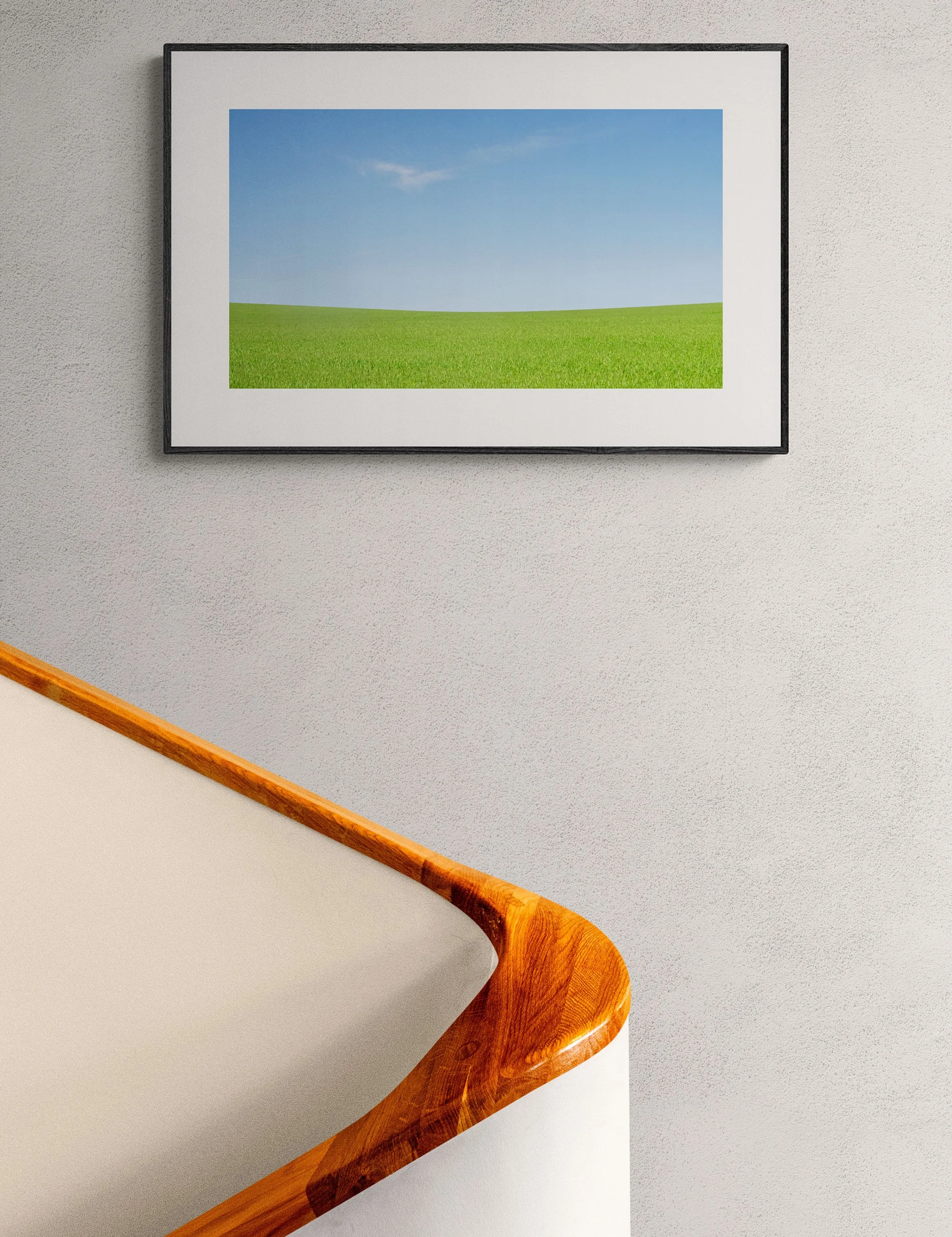 “Cloud Cradle” framed minimalist landscape print by Ian J. Muirhead Arts above a curved timber staircase in a minimalist interior. A sweeping green field beneath an open blue sky with wisp of cloud..