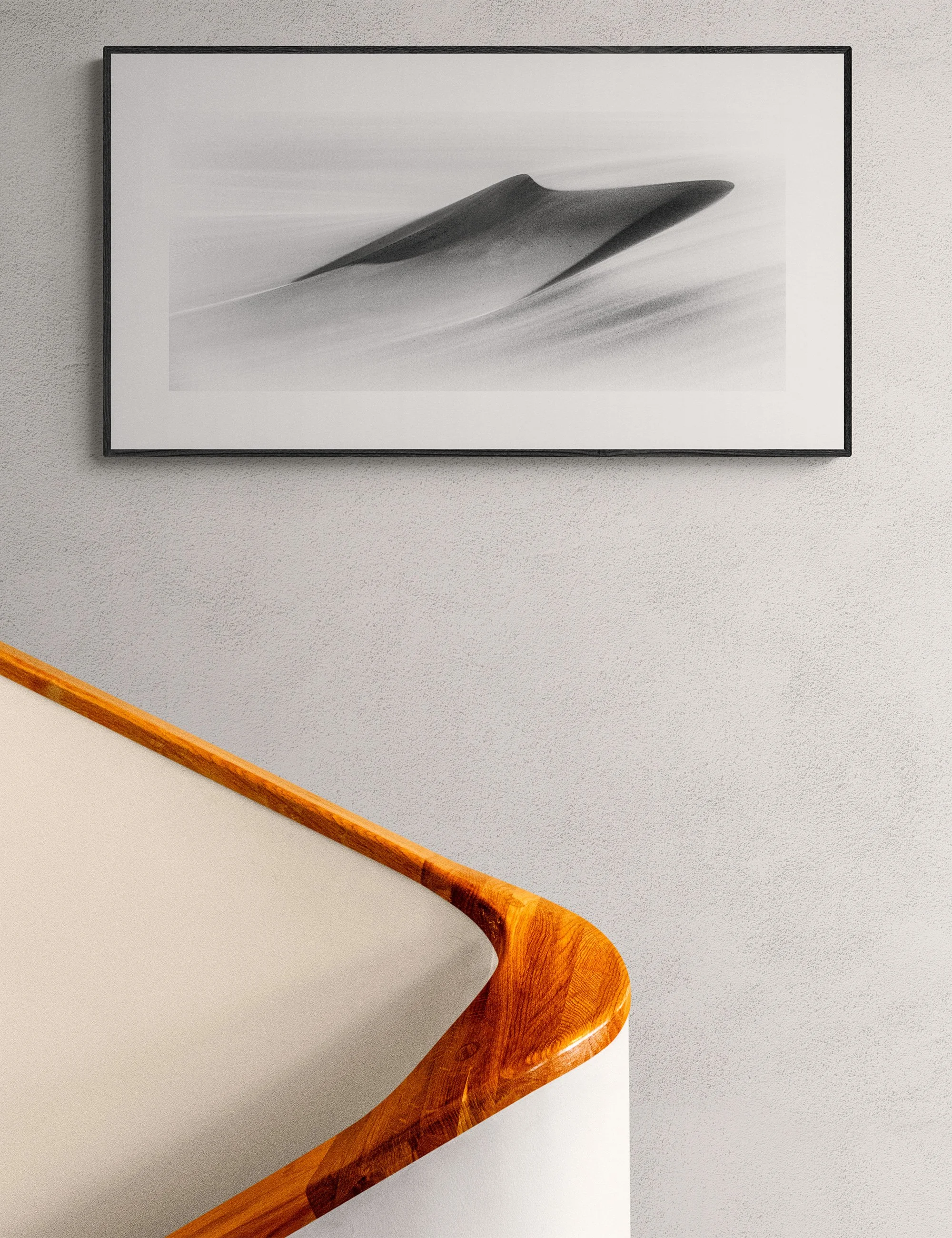 “A Harsh Land” framed minimalist landscape print by Ian J. Muirhead Arts above a curved timber staircase in a minimalist interior. A lone tree and sweeping tracks create a stark rural scene.