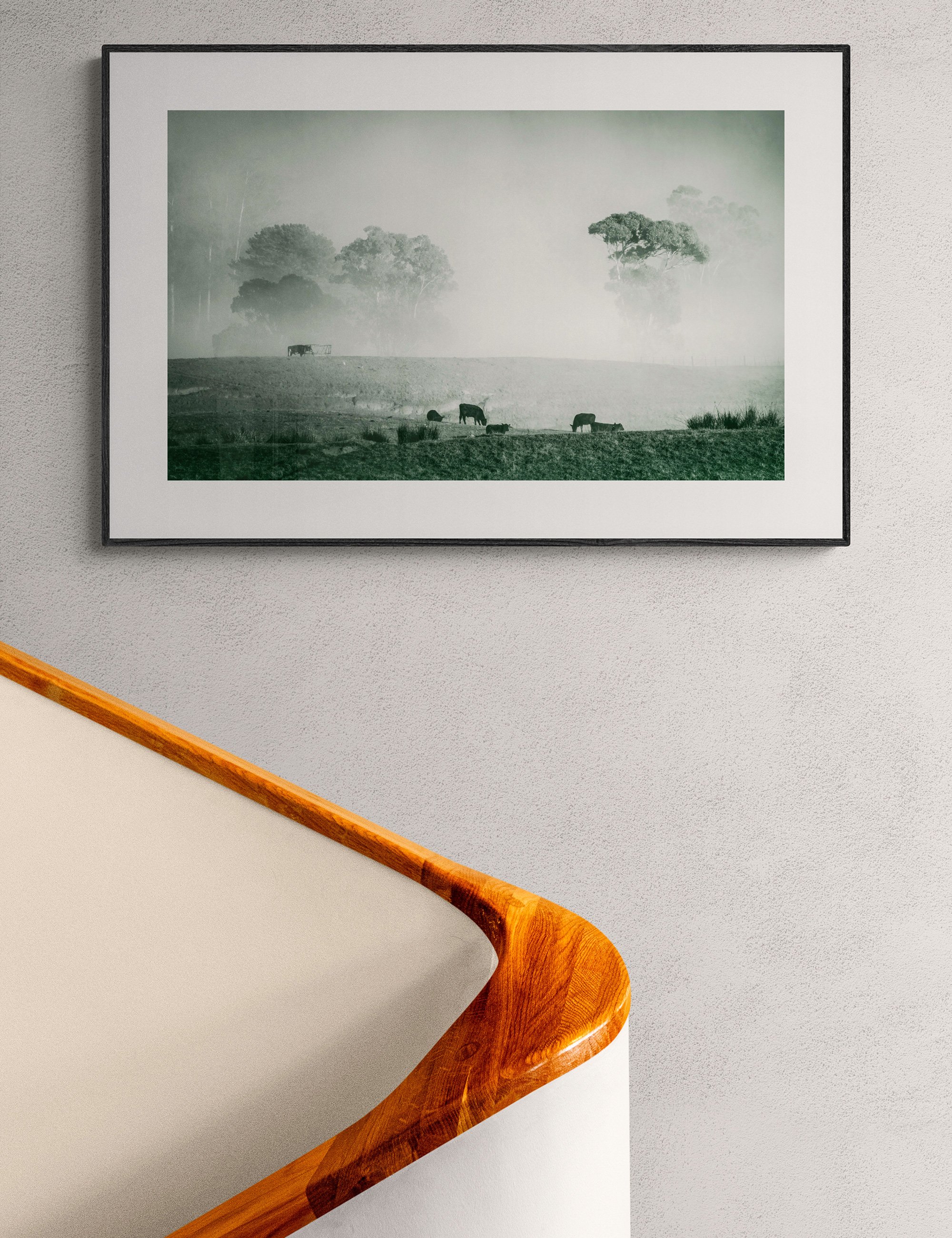 “Pastoral Calm” framed landscape print by Ian J. Muirhead Arts above a curved timber staircase in a minimalist interior. Grazing cattle and misty trees create a quiet rural atmosphere.