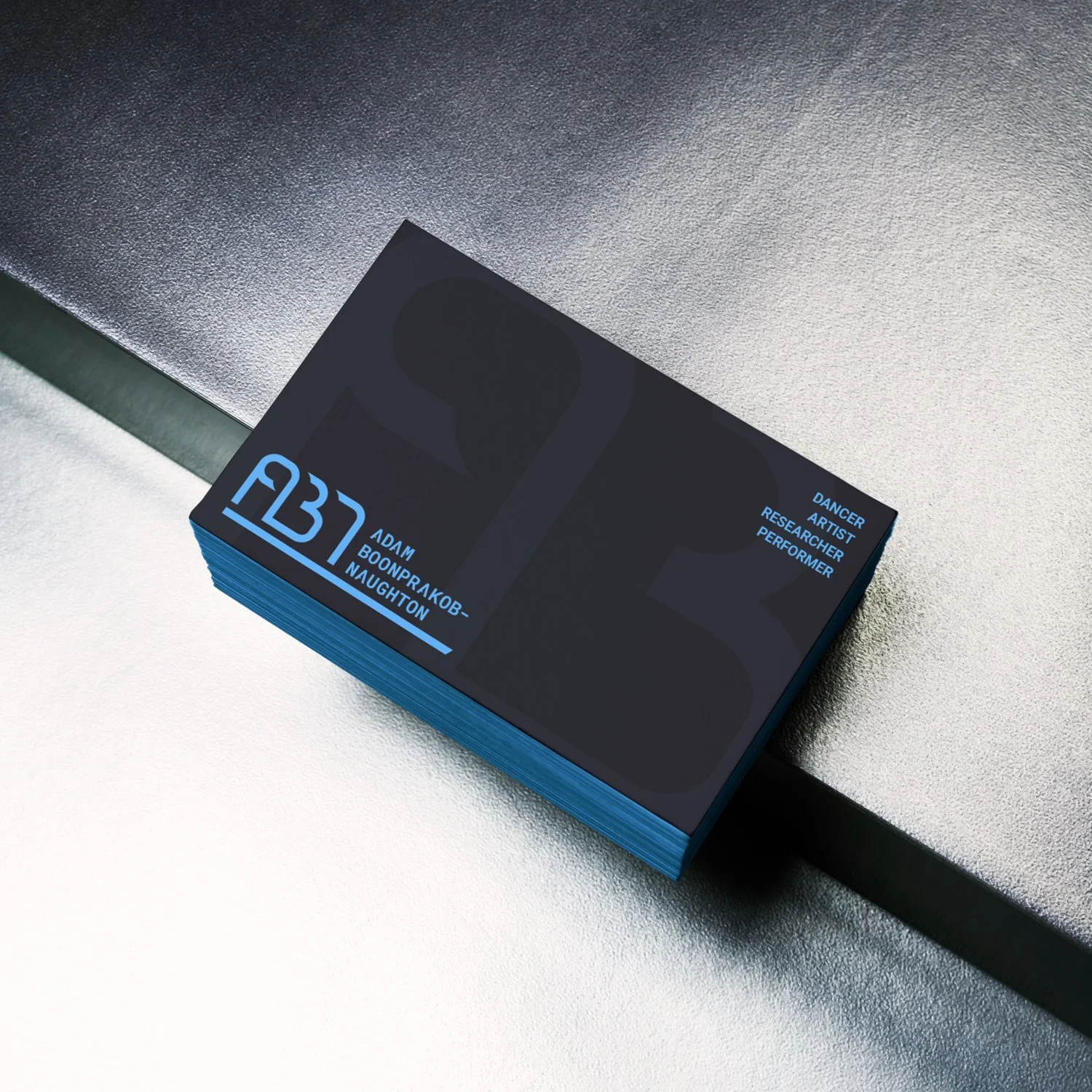 A set of business cards with a blue and black design, featuring a large logo and text about a person named Adam Booonprakob, who is a dancer, artist, researcher, and performer, placed on a metallic surface.