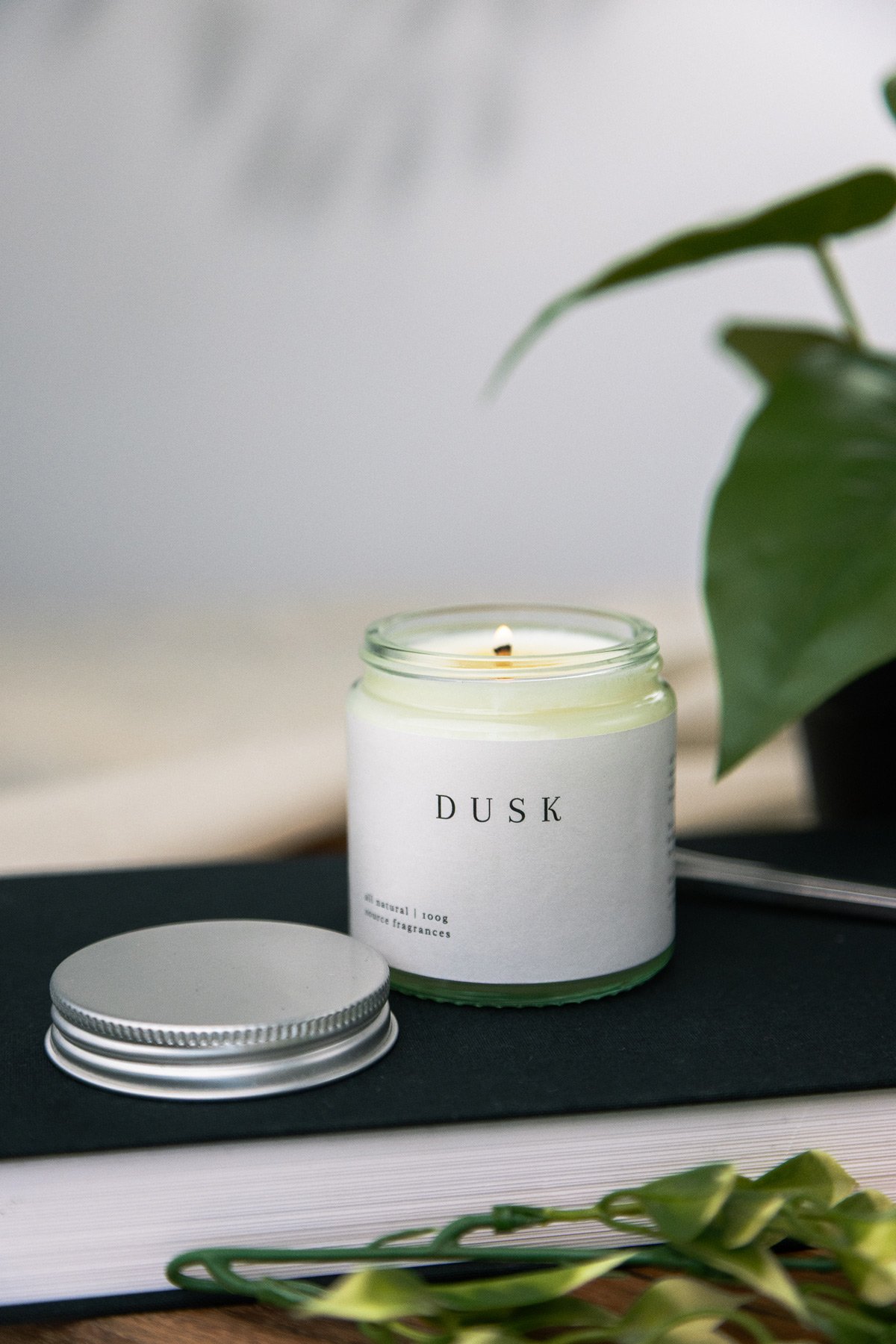 A lit white candle in a glass jar labeled 'DUSK' resting on a black surface next to a closed metal lid, with a plant with large green leaves partially visible on the right.