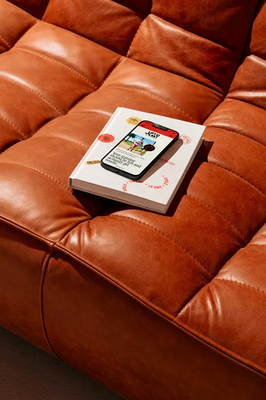 A book with a phone on top of it resting on a brown leather couch.