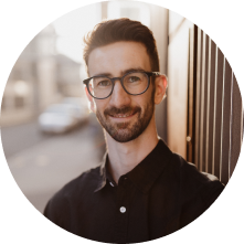 A young man with glasses and a beard smiling outdoors near a wooden fence, with a blurred street in the background.