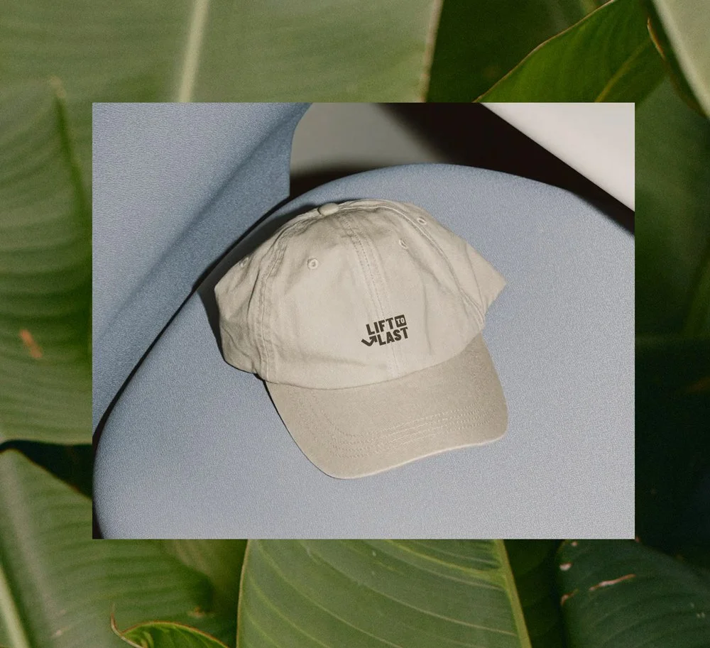 Beige baseball cap with the text 'Lift to Last' on the front, placed on a blue surface with green leaves in the background.