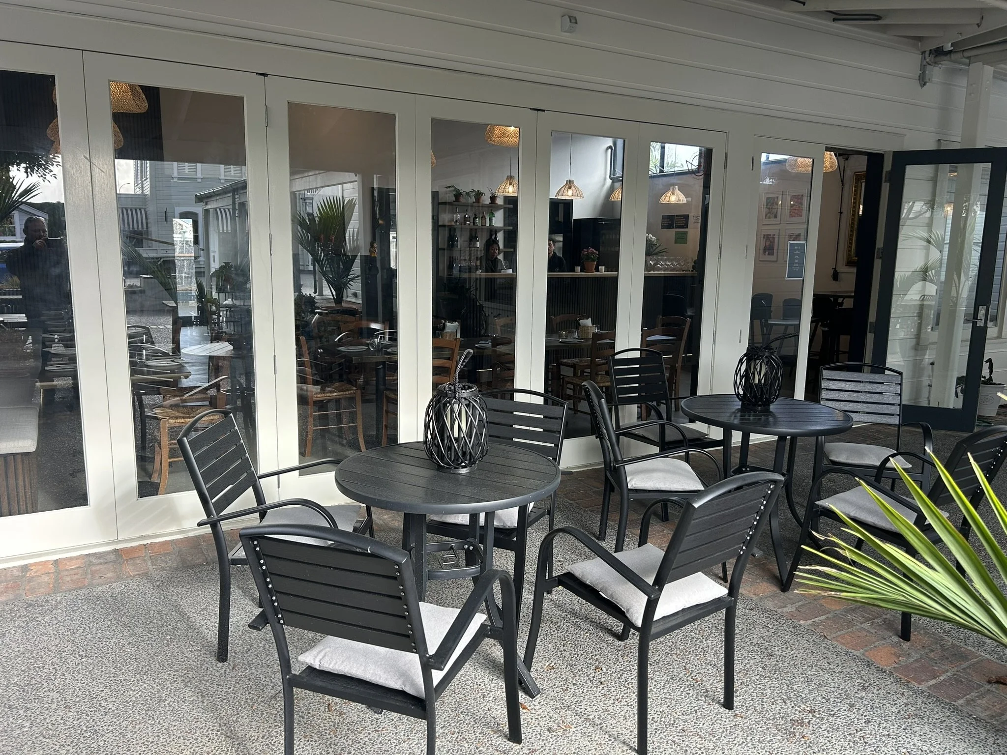 Outdoor seating area with black metal tables and chairs, visible through large glass windows of a cafe or restaurant, with pendant lights inside and some greenery visible outside.