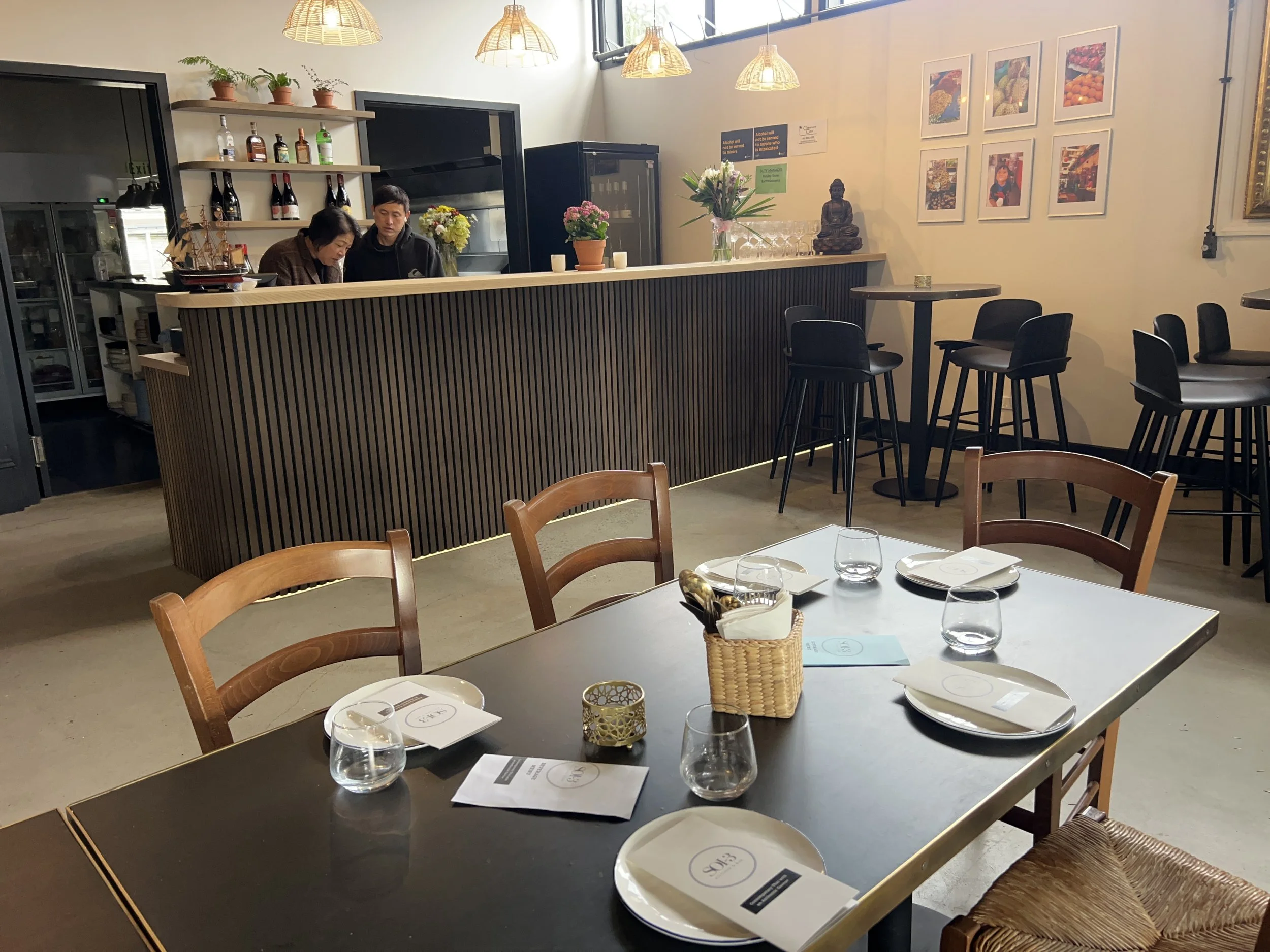 Interior of a modern restaurant with high ceilings and warm lighting. A bar counter with two people, a woman and a man, is visible behind it. On the wall to the right, there are framed pictures of colorful fruits and vegetables. In the foreground, a 
