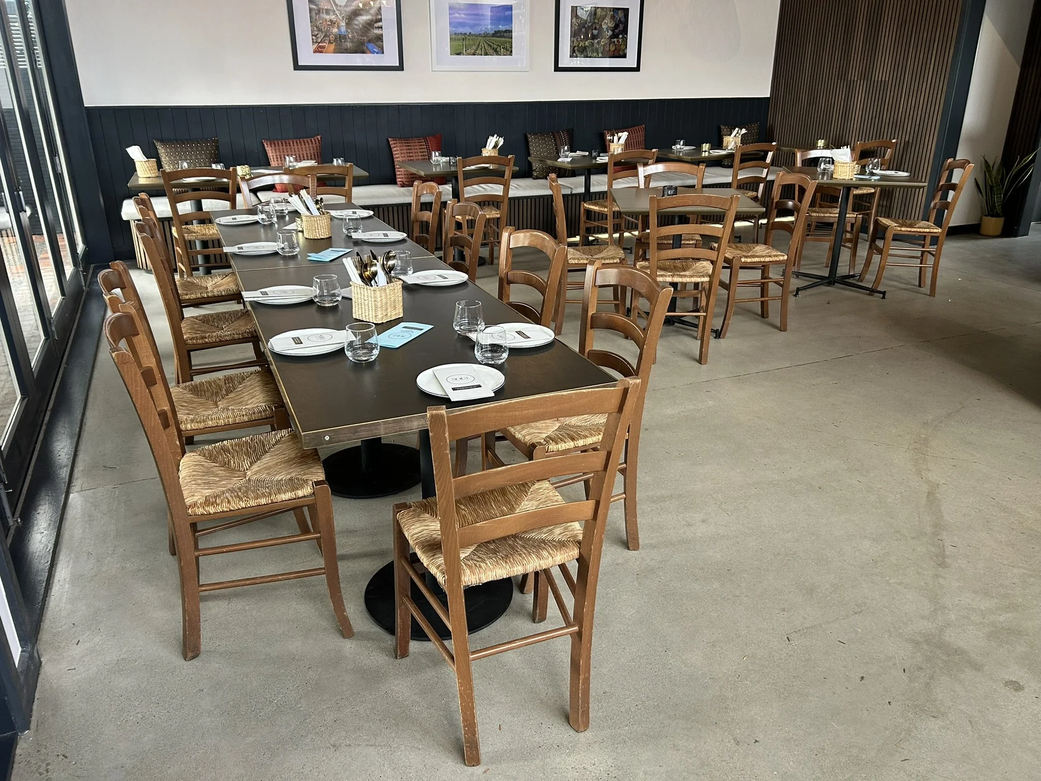 Interior of a restaurant with wooden tables and chairs, set with glasses, plates, and utensils, decorated with pillows along the wall, with framed photographs on the wall and a potted plant in the corner.