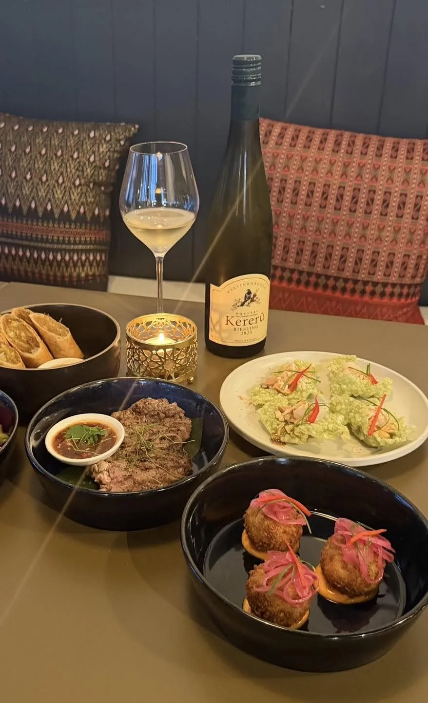 A table with a glass of white wine, a bottle of Kereru Riesling, a small candle, and various appetizers including croquettes, a plate of lettuce wraps, grilled meat with dipping sauce, and a dish with fried bites topped with pink garnish.