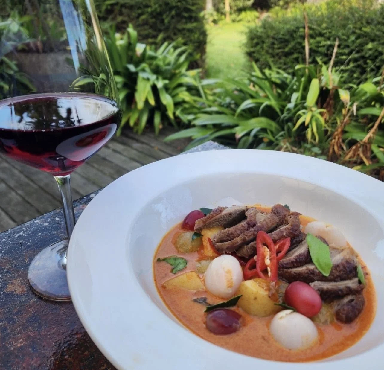 A glass of red wine and a plate of stew with vegetables outdoors on a wooden table in a garden.