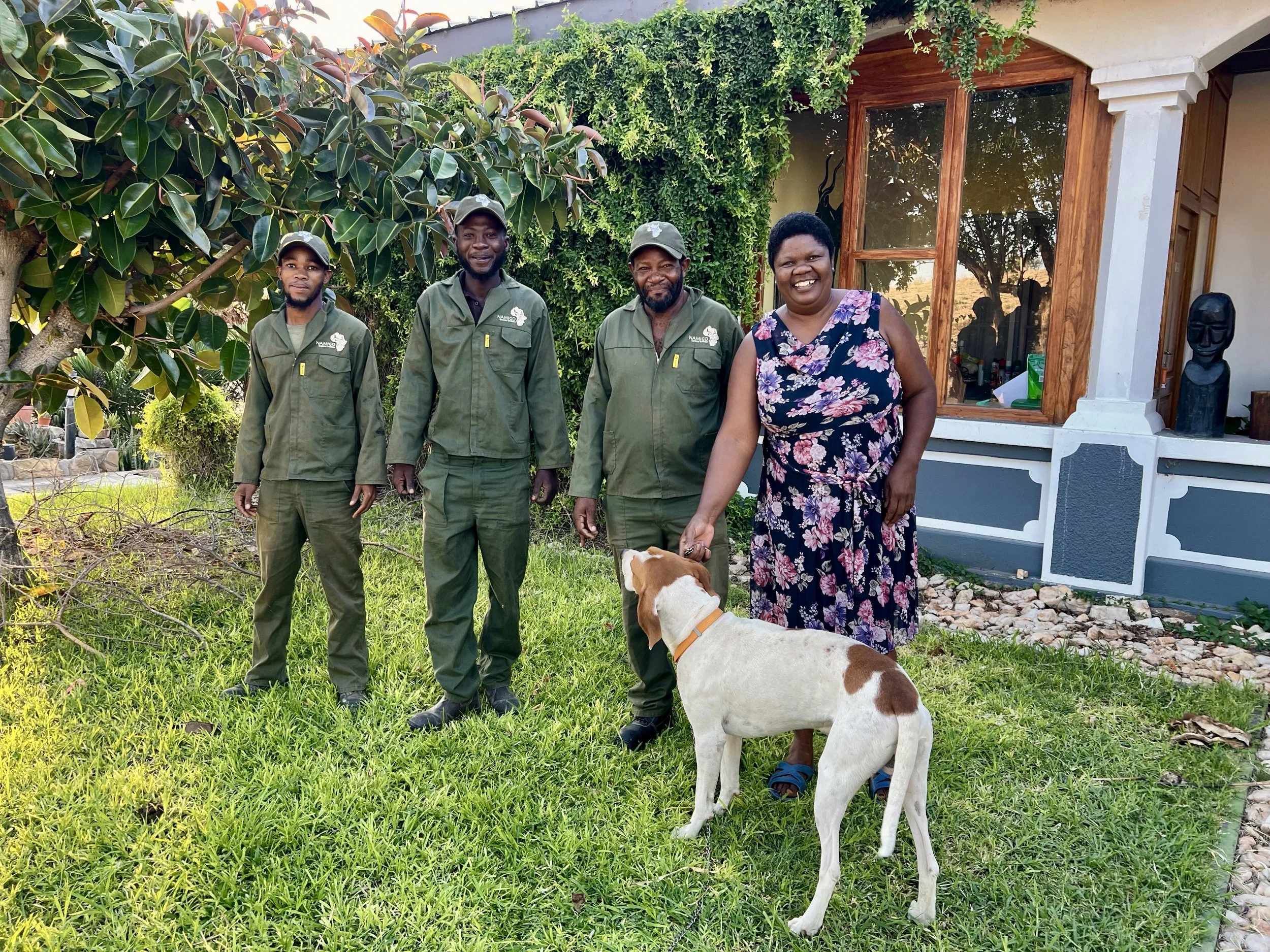 Vier Männer stehen vor einem Tor, umgeben von Pflanzen und Blüten, während sie in die Kamera lächeln.