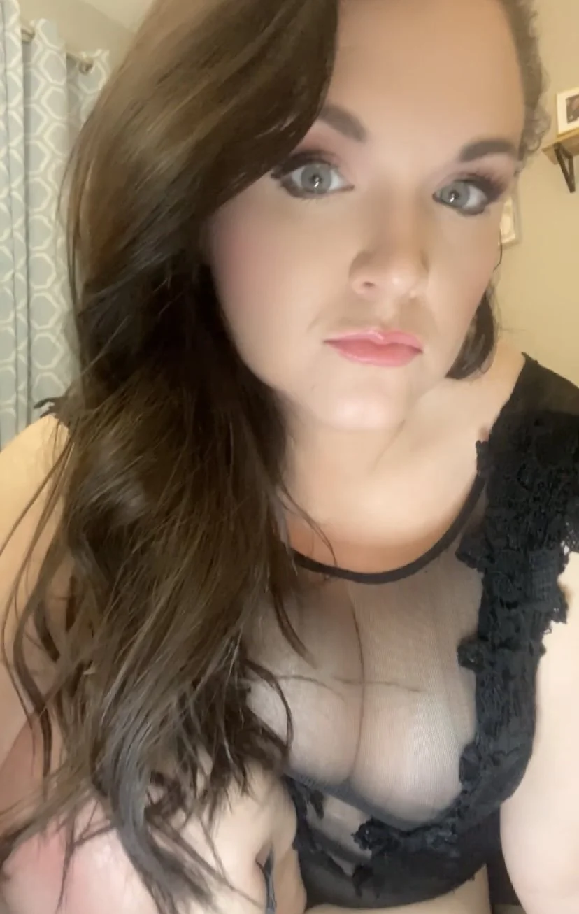A woman with long wavy brown hair and blue eyes wearing a black lace top with sheer fabric.