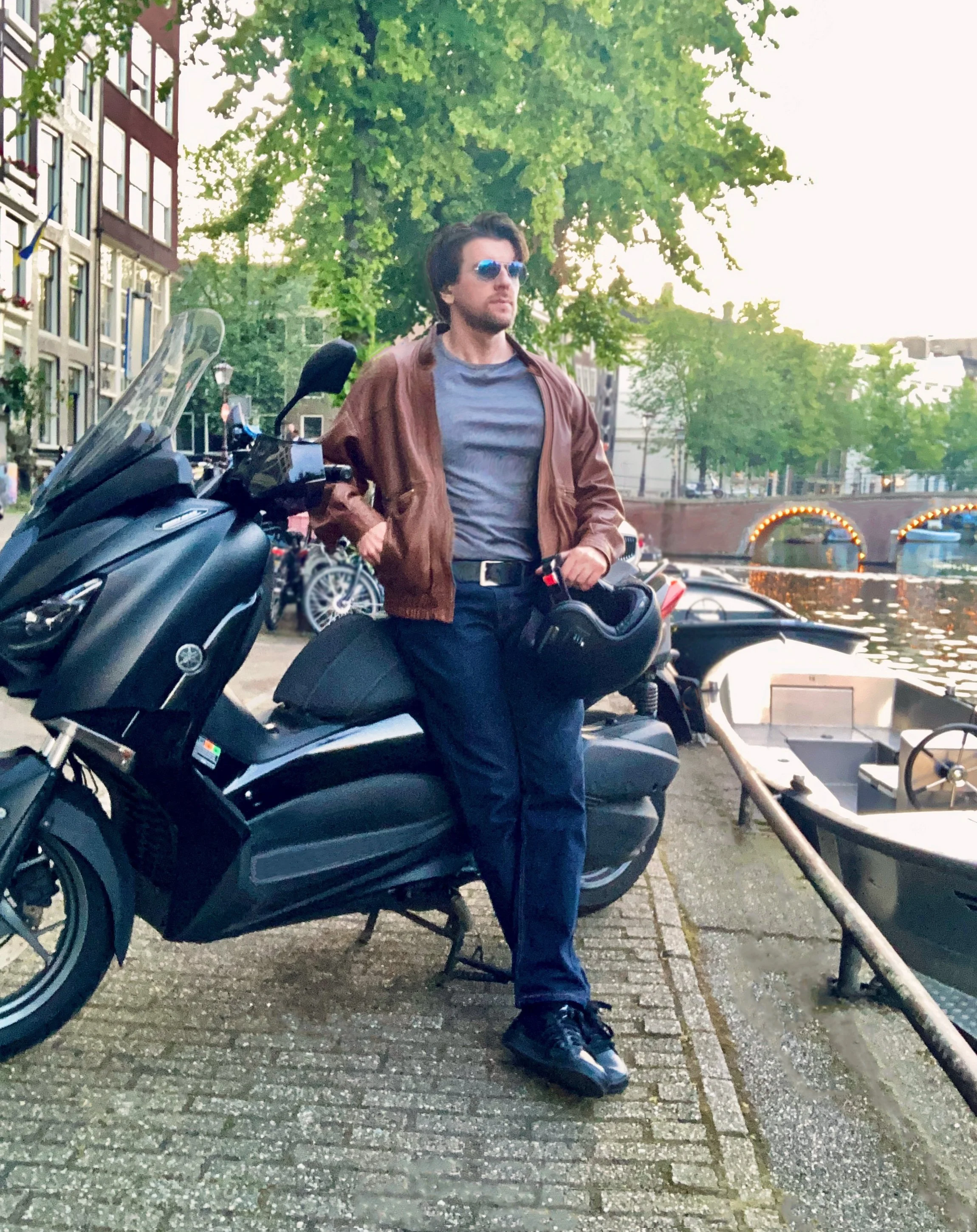A man with dark hair, wearing sunglasses, a brown leather jacket, gray t-shirt, black jeans, and black sneakers, stands beside a black scooter next to a canal with boats and lit arches in the background.