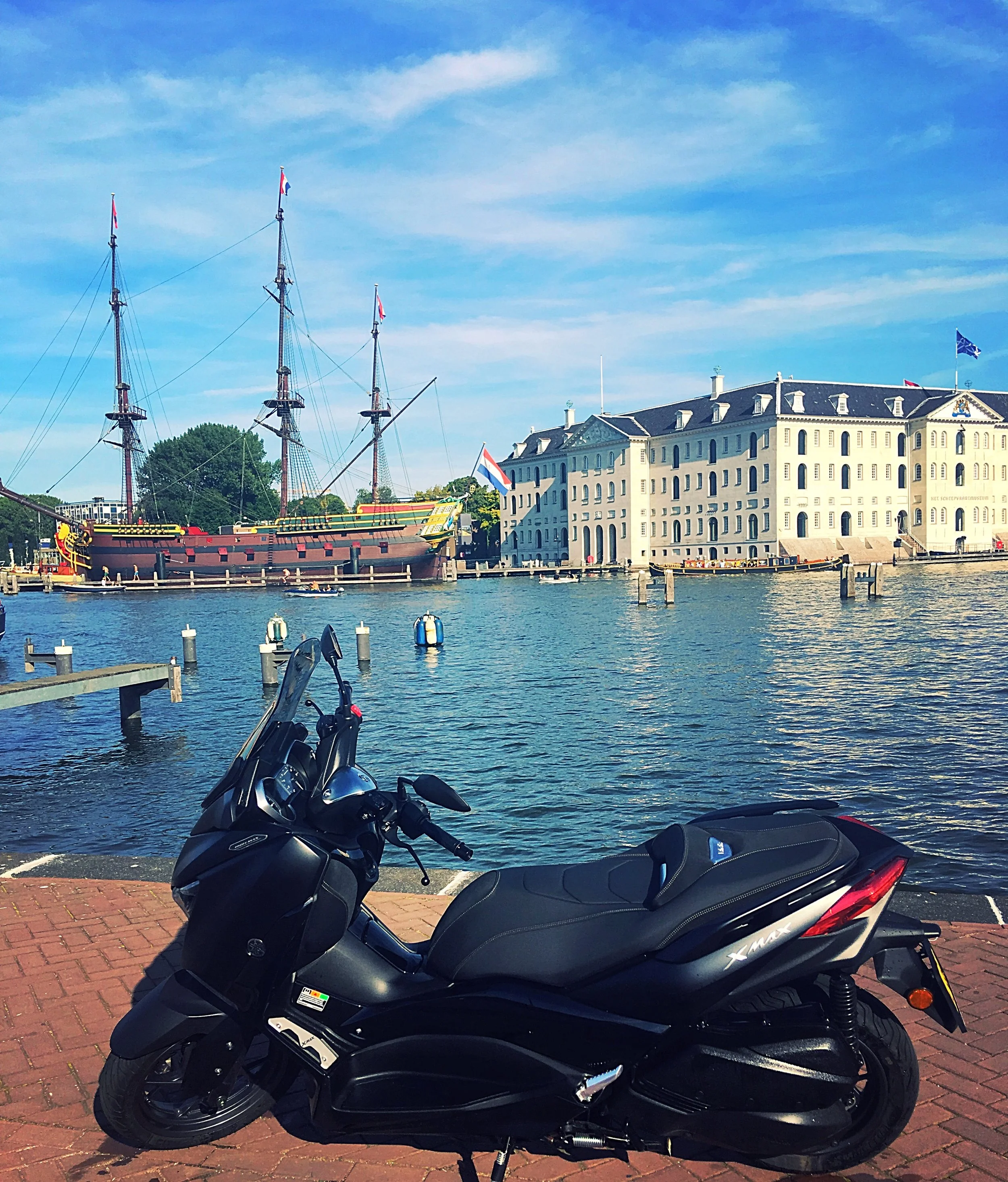 Black scooter parked on brick sidewalk near water with historic ship and large white building in the background under a blue sky.