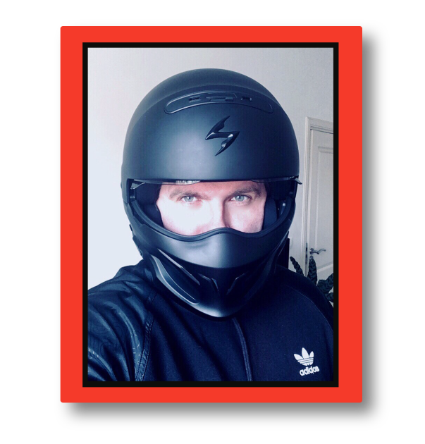 Person wearing a black motorcycle helmet and black Adidas jacket, taking a selfie with a backdrop of a white wall and a closed door.