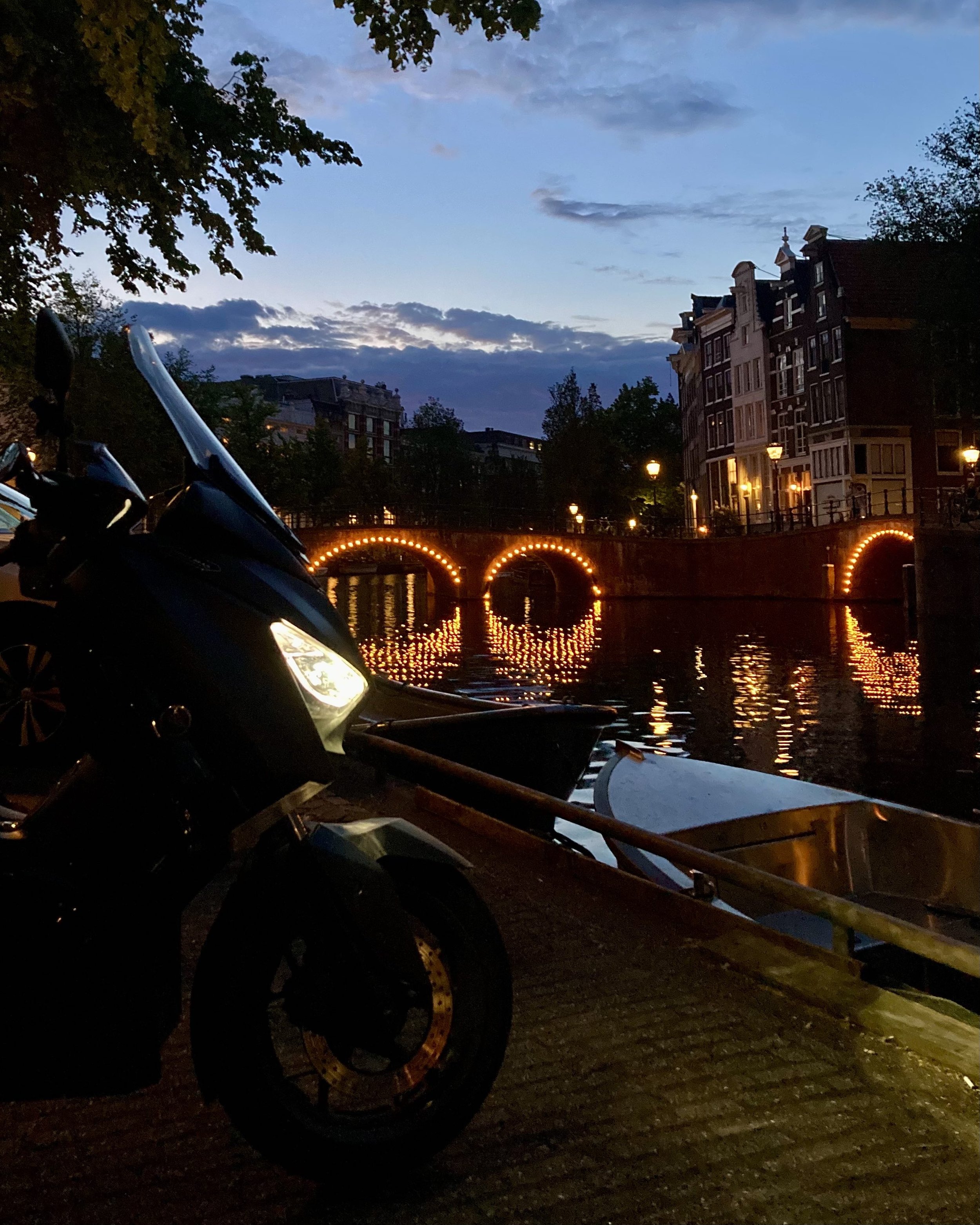 A nighttime scene of a river with a bridge decorated with orange lights, reflecting in the water. On the left, a black motorcycle is parked beside a boat.