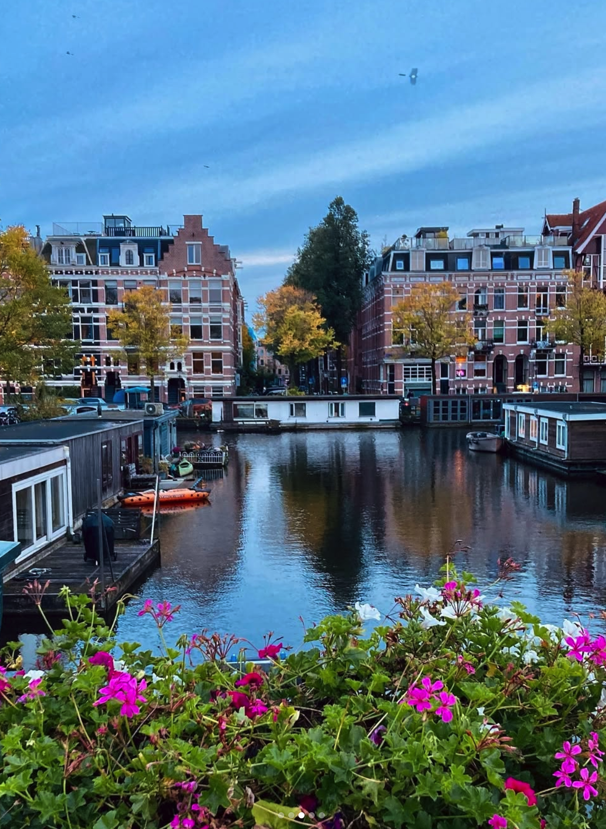 View of a canal with houseboats and colorful flowers in the foreground, historic buildings with gabled roofs, trees with autumn foliage, and a small airplane flying in a partly cloudy sky.