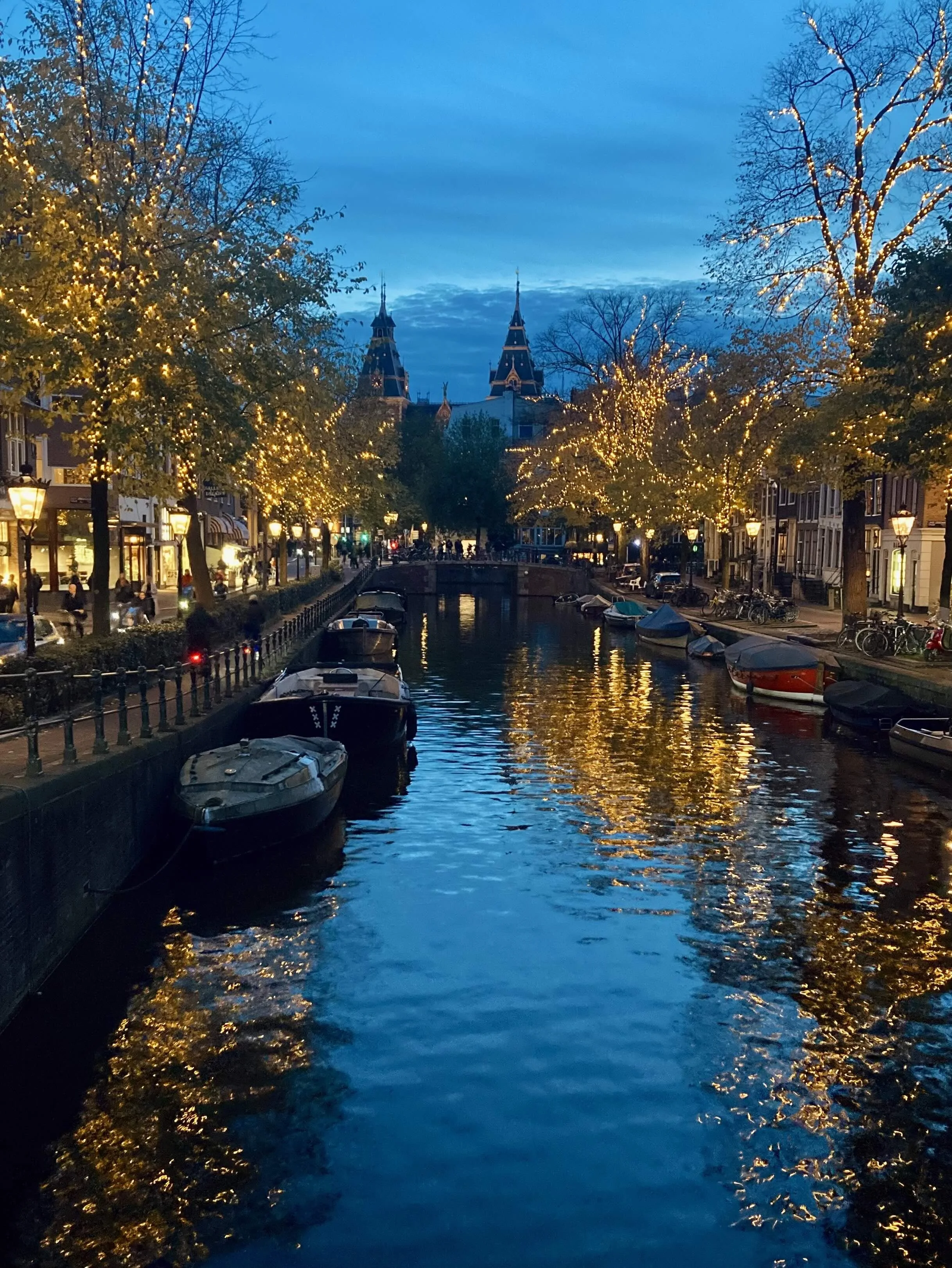 A city canal at dusk with boats moored along the sides, illuminated trees with string lights, historic buildings, and a church with twin spires in the background.