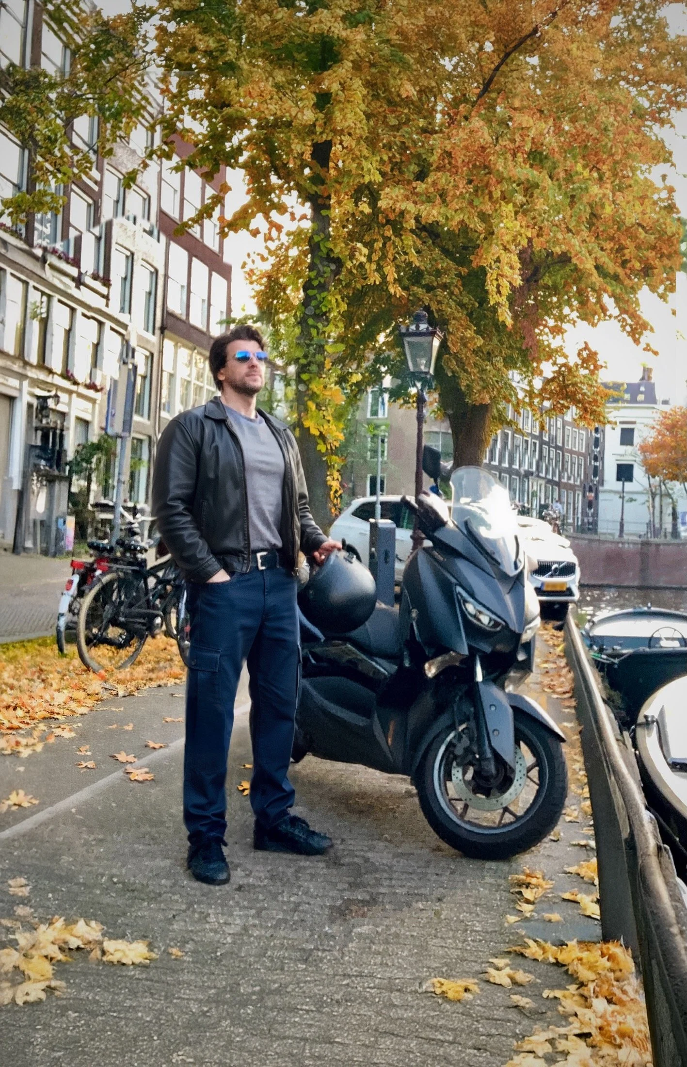 A man wearing sunglasses and a black leather jacket standing next to a black scooter on a city street during autumn, with fallen leaves on the ground and colorful trees in the background.