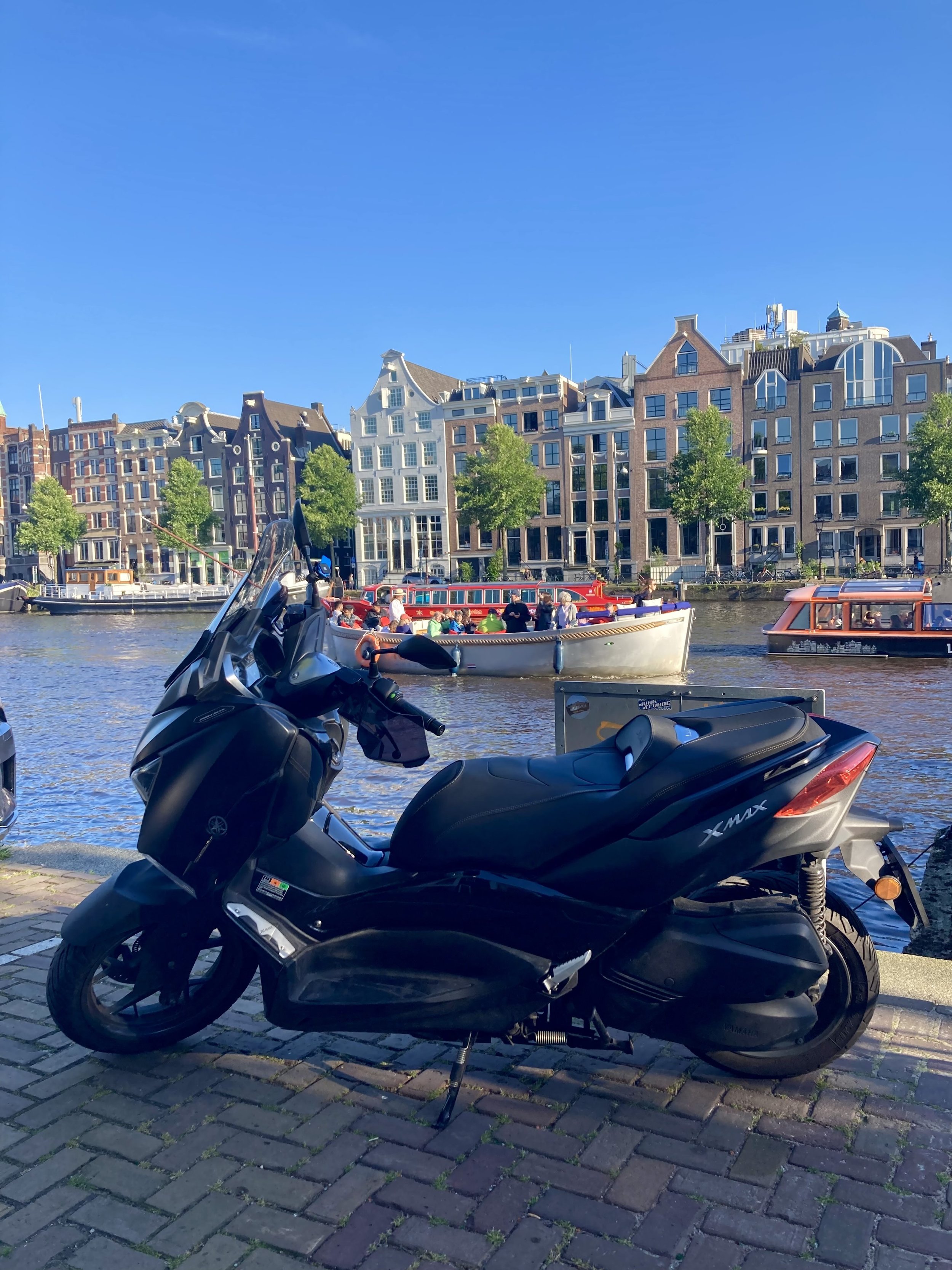 A black Yamaha XMAX scooter parked on a brick sidewalk near a canal with boats and colorful buildings in the background under a clear blue sky.