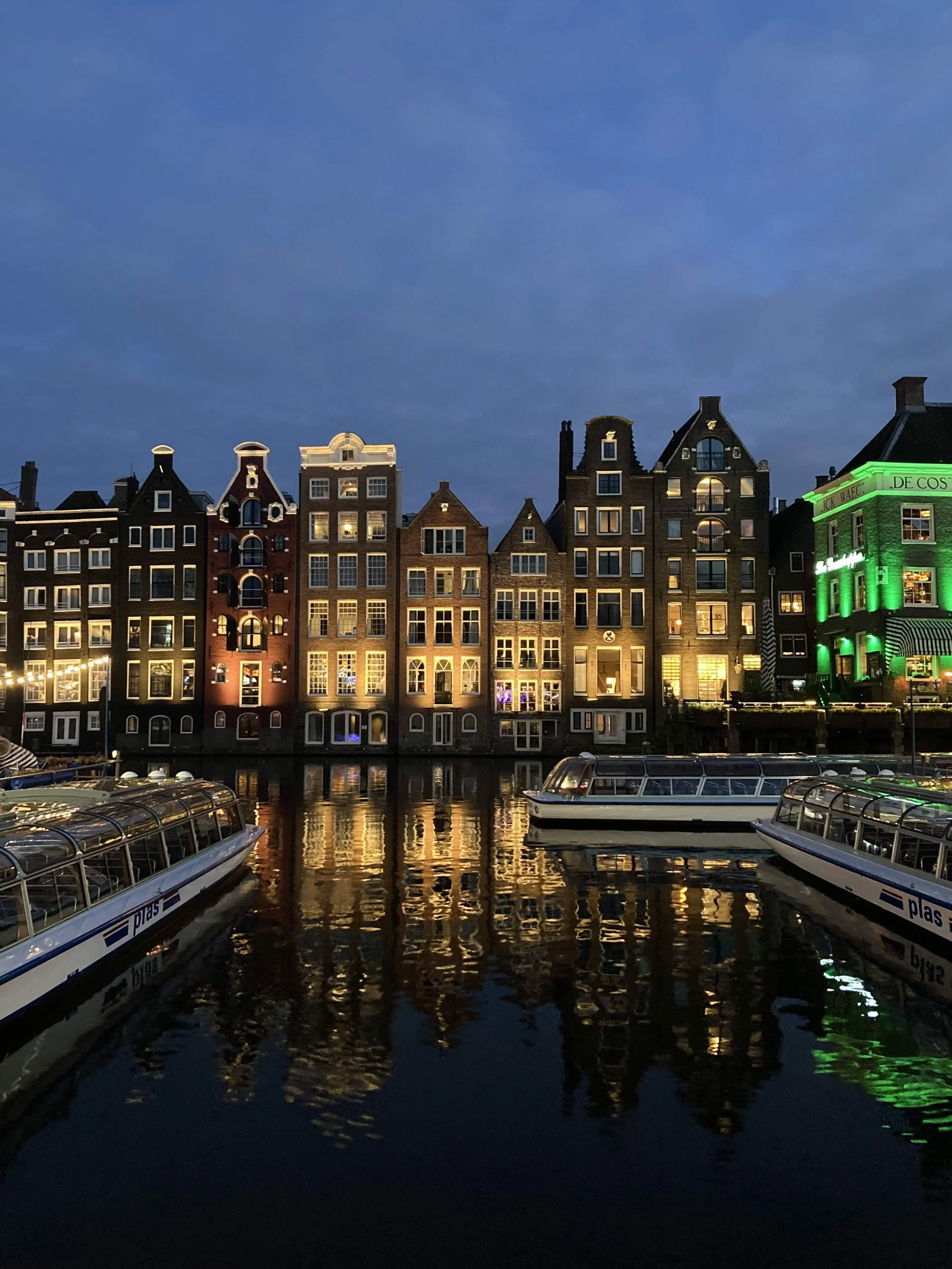 Nighttime view of colorful buildings along a canal with reflections in the water and boats docked nearby.