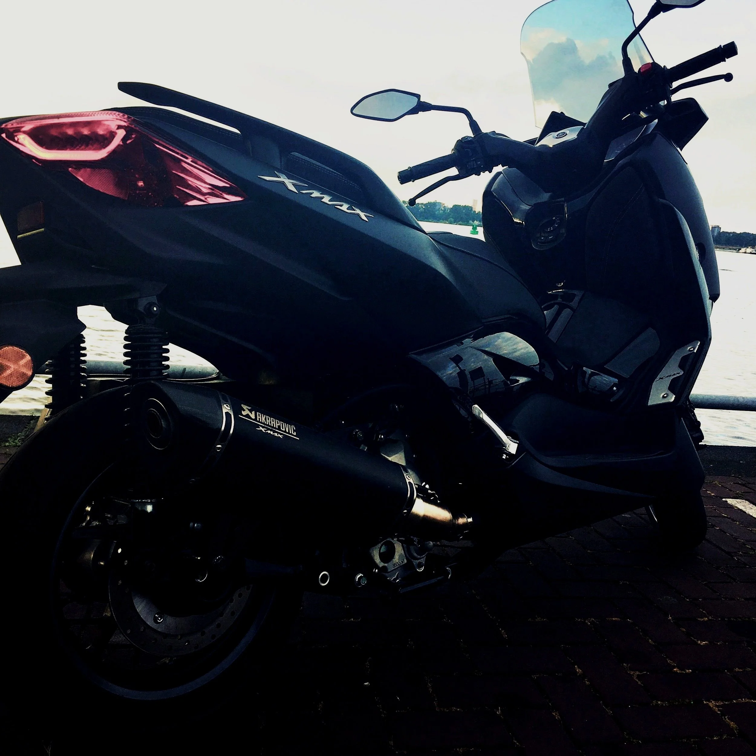 A black Yamaha XMAX scooter parked near a body of water, with a bridge and a cloudy sky in the background.