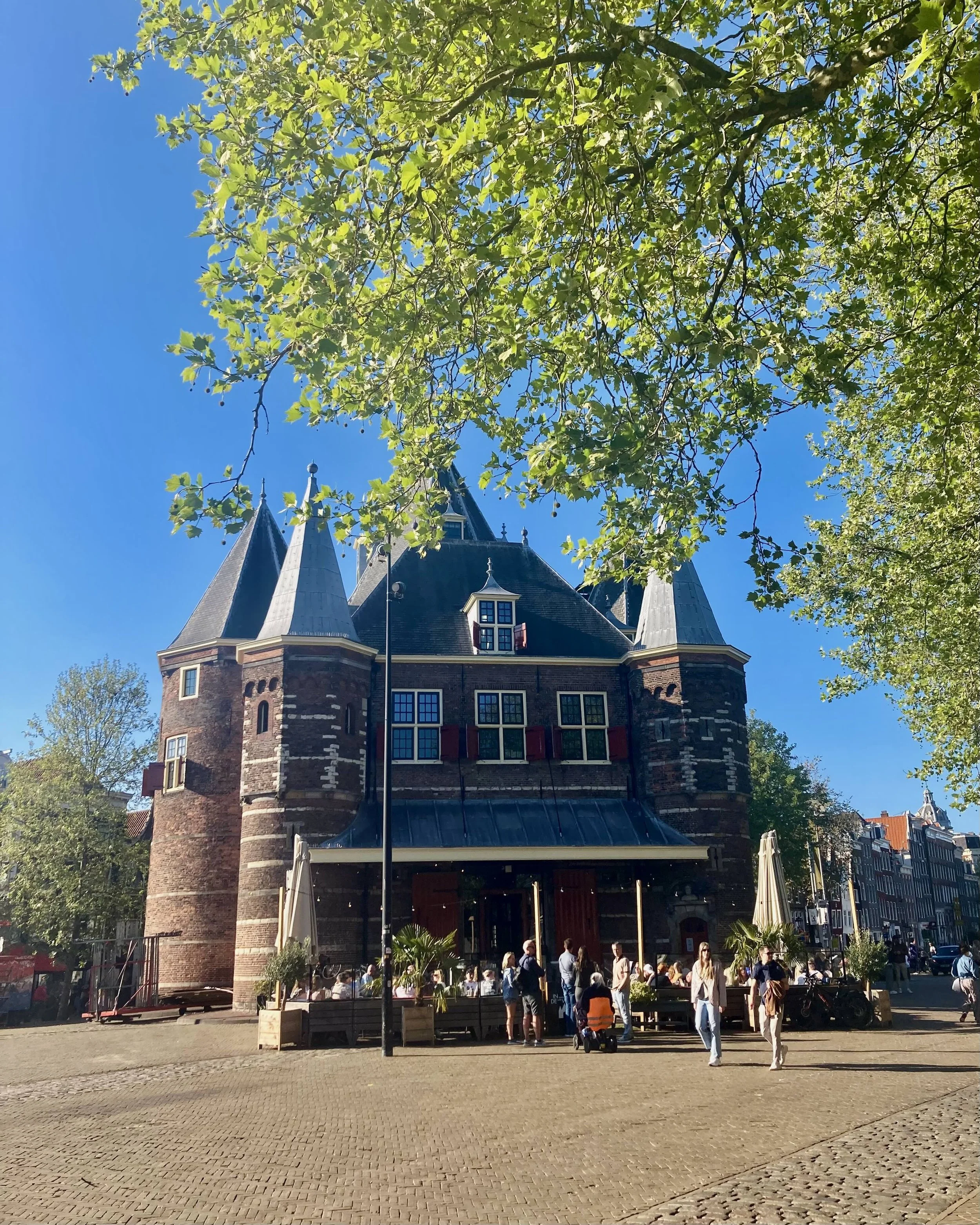 A historic brick castle-like building with two prominent towers and a peaked roof, located on a busy street with people walking nearby, framed by green tree branches against a bright blue sky.