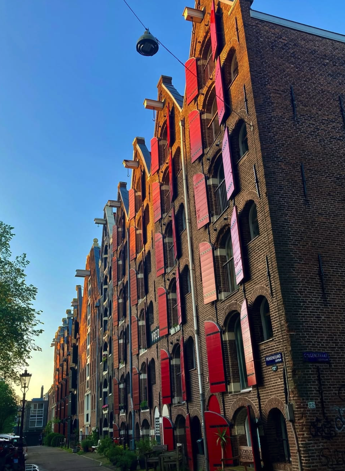 A multi-story brick building with red and black window shutters, street signs, and a street lamp, taken during daylight.