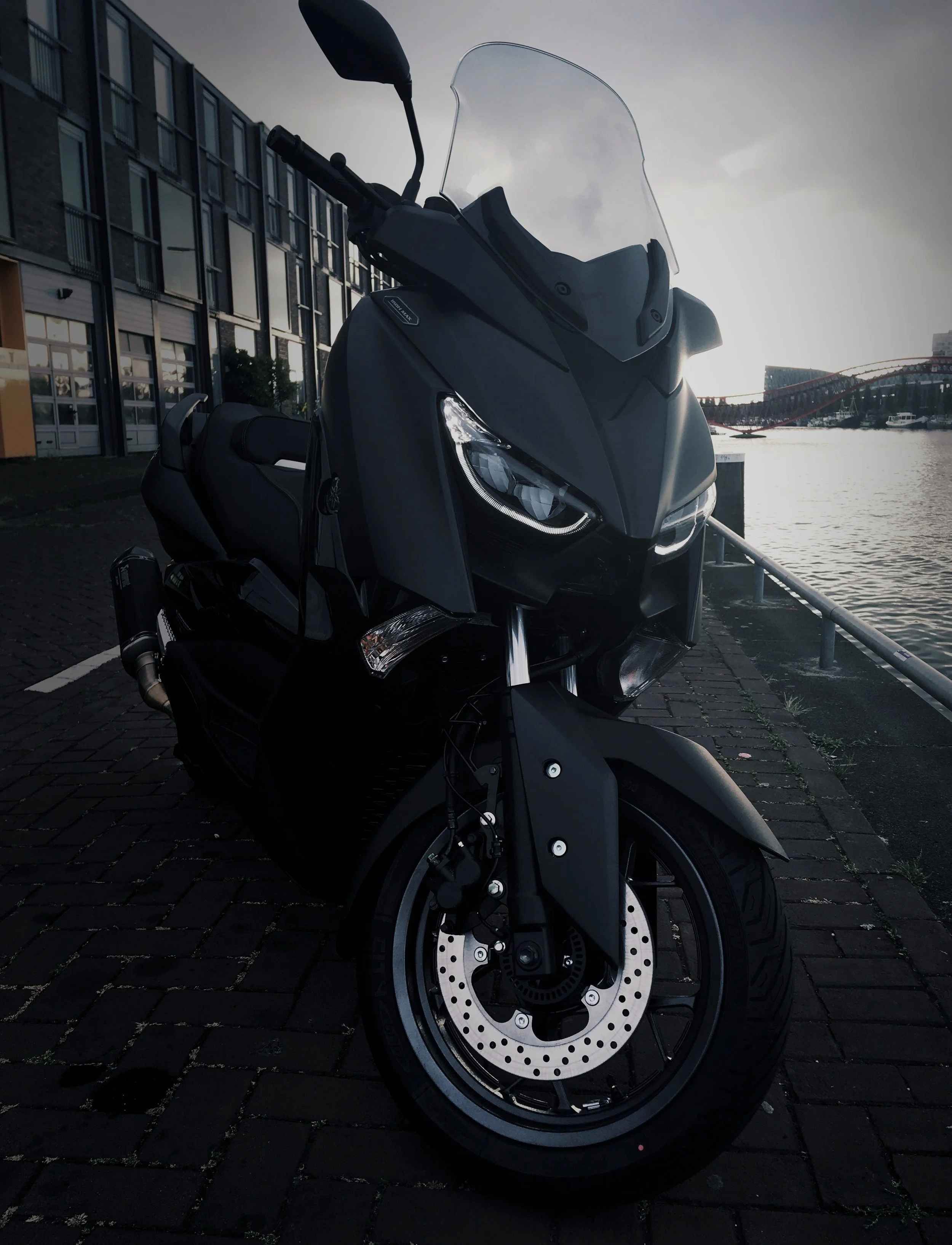 Black touring motorcycle parked on a brick pavement near a body of water with modern buildings and a bridge in the background at sunset.