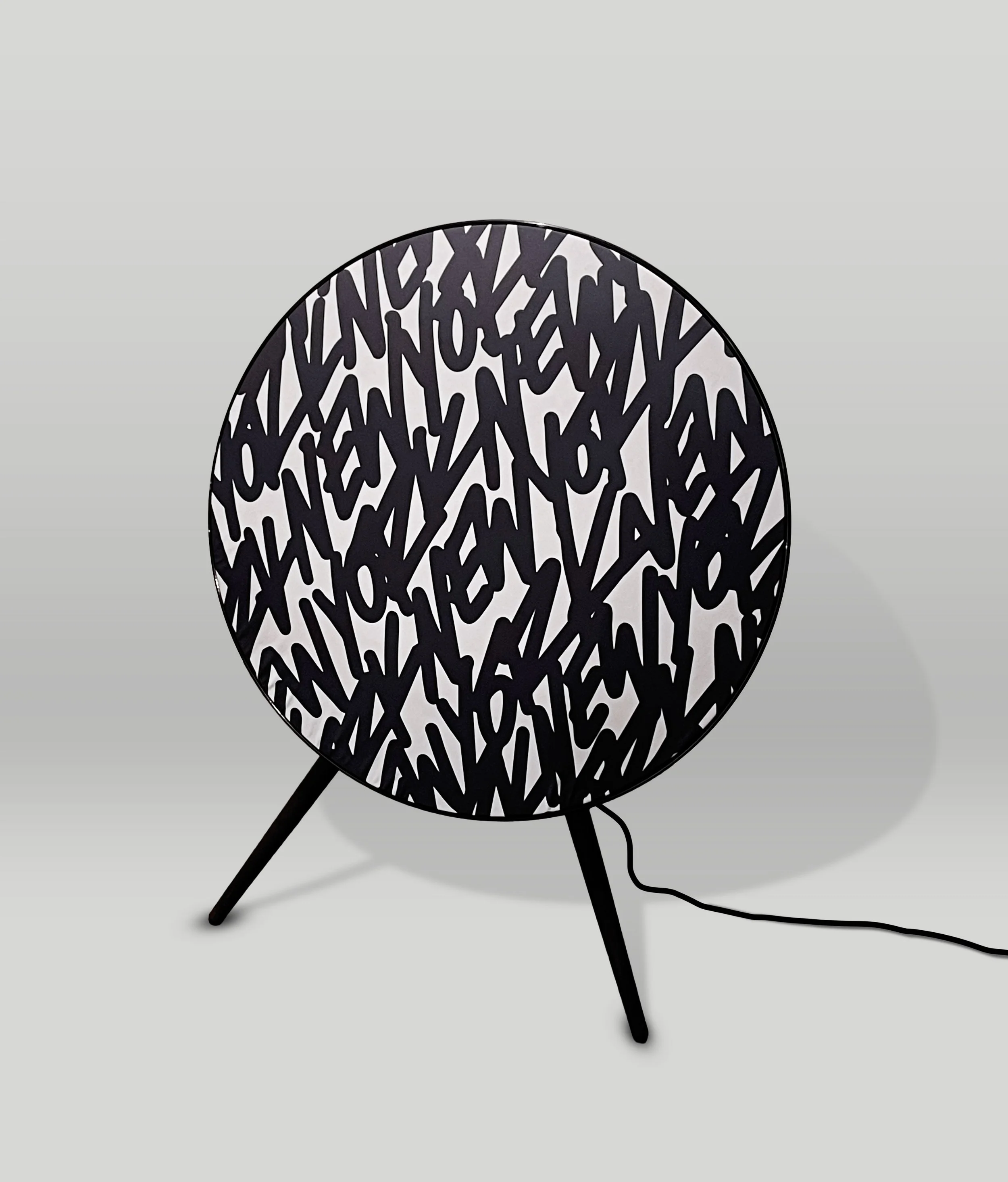 Ein schwarzer Lüster mit einer kunstvollen, graphischen Musterung aus schwarzen Linien auf weißem Hintergrund, steht auf einem grau gefärbten Boden. Es ist ein modernes Beleuchtungselement mit Metallrahmen und Kabel, das Schatten auf dem Boden wirft.
