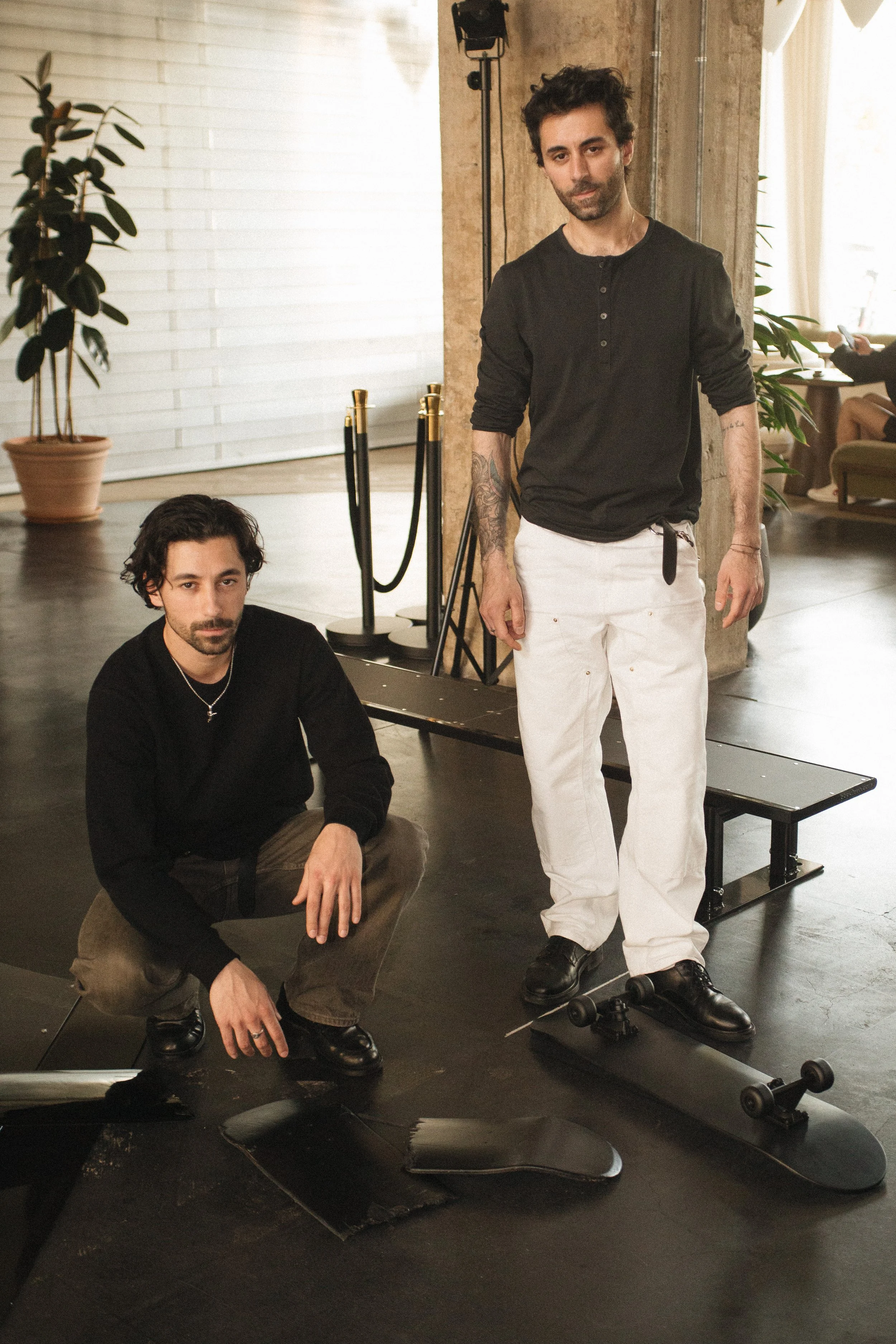 Zwei Männer, einer sitzt auf dem Boden und der andere steht neben einem Skateboard, in einem modernen Raum mit Pflanzen und Beleuchtung.