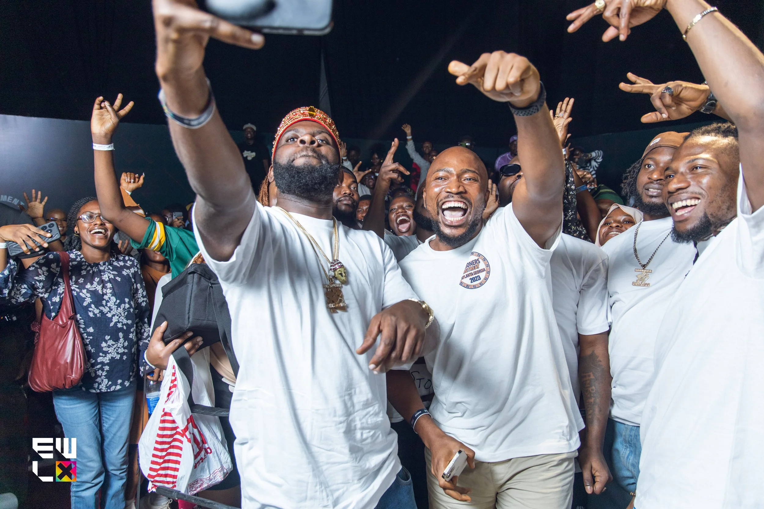 A group of people celebrating outdoors, smiling and making gestures, with some wearing white shirts and accessories, at night under dark sky.
