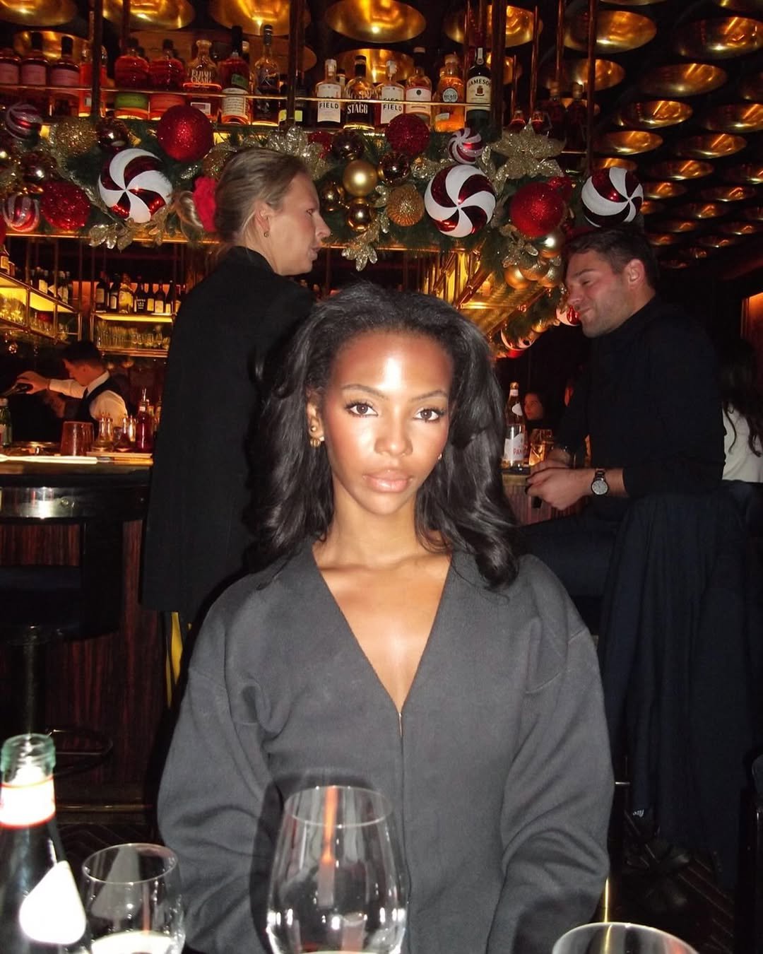 A woman with dark hair and a gray zip-up top sitting at a restaurant table with wine glasses and bottles in front of her, decorated with Christmas ornaments and garland, with two people working or socializing in the background.