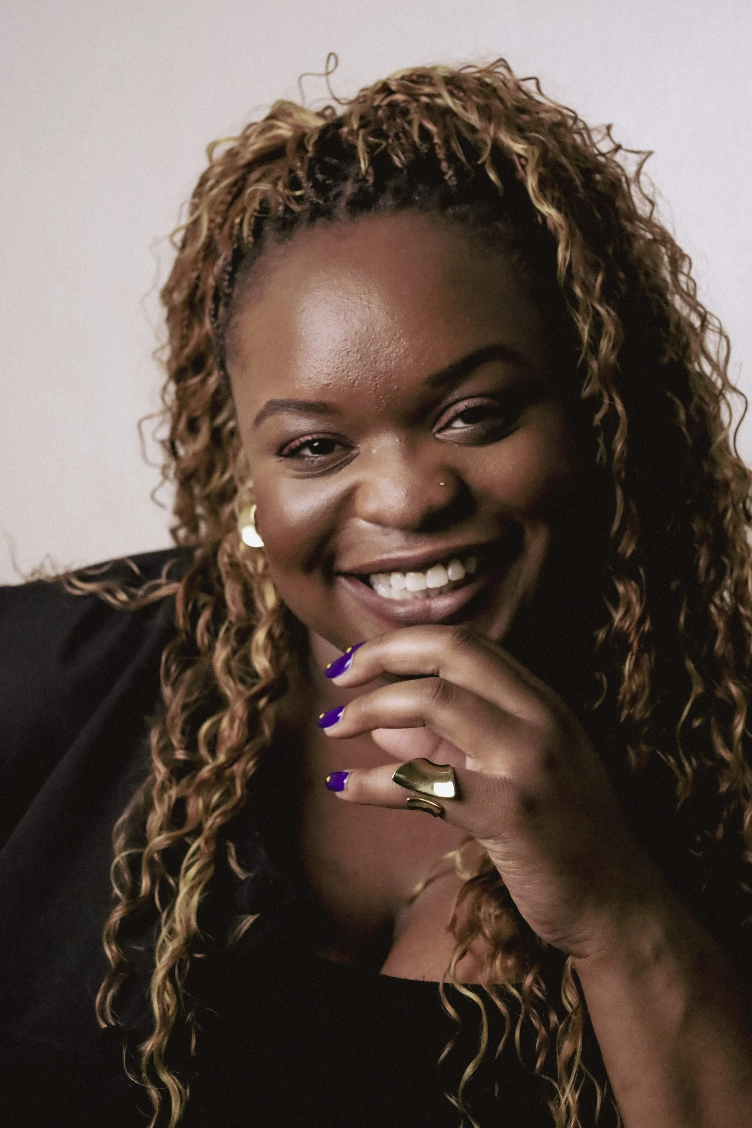 Close-up of a smiling woman with curly, blonde-tinted hair, wearing gold earrings, a gold ring on her finger, and a black top, with dark purple nail polish.