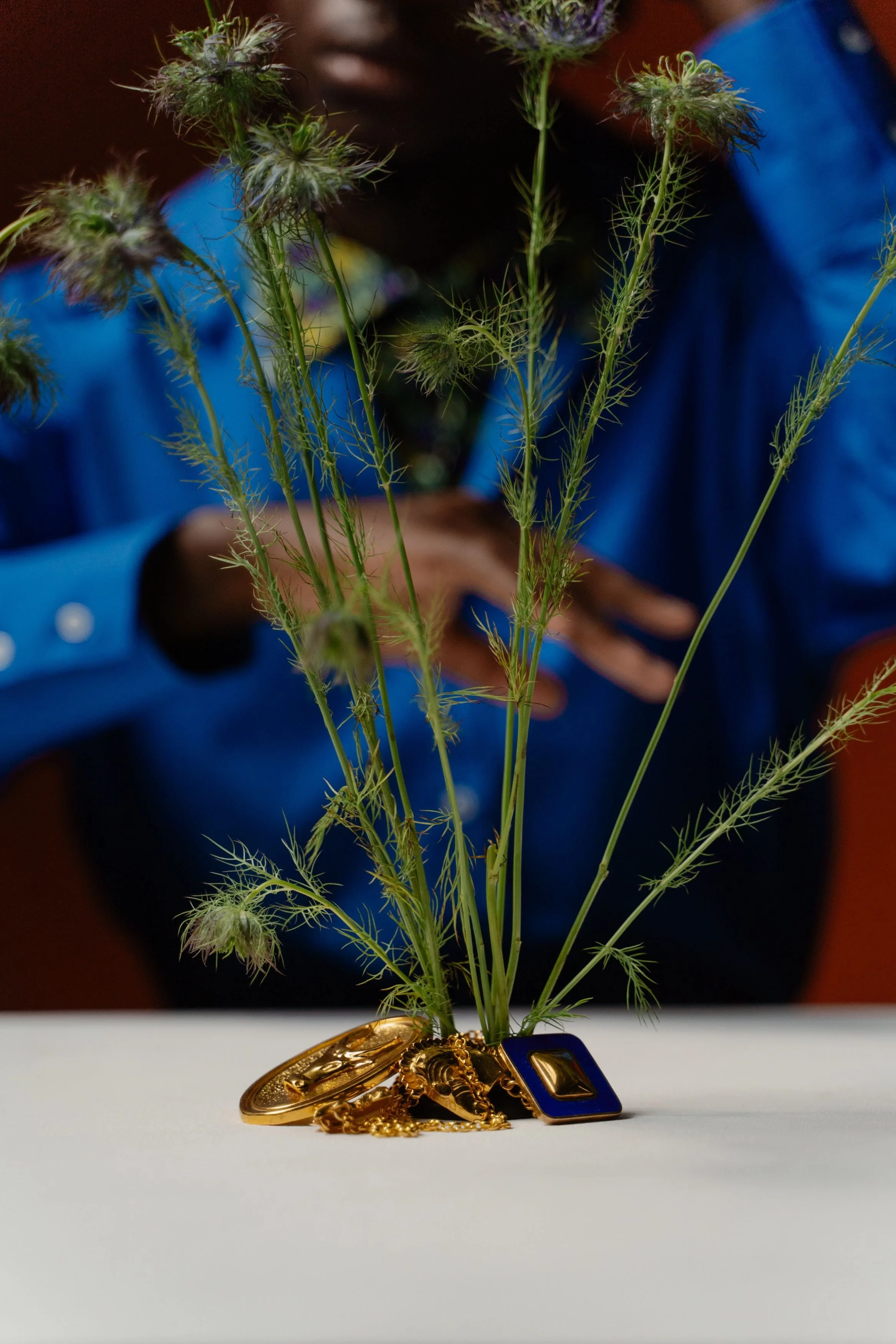 A small plant with green, feathery leaves is placed in a pot decorated with gold chains and a gold watch, on a white surface, with a blurred person wearing a blue shirt in the background.