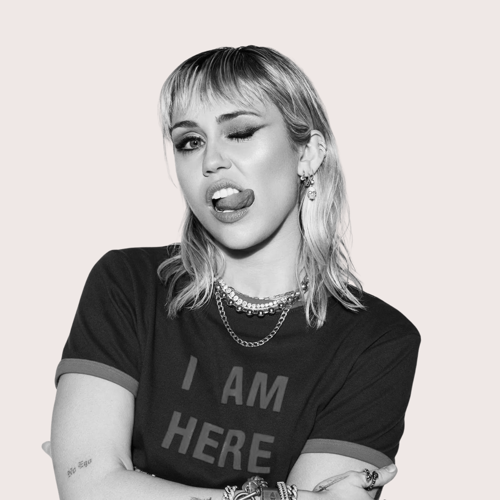 A woman making a playful face with her tongue out and one eye closed, wearing a black t-shirt that says 'I AM HERE,' multiple necklaces, earrings, and bracelets, against a plain background. Women’s confidence empowerment training course