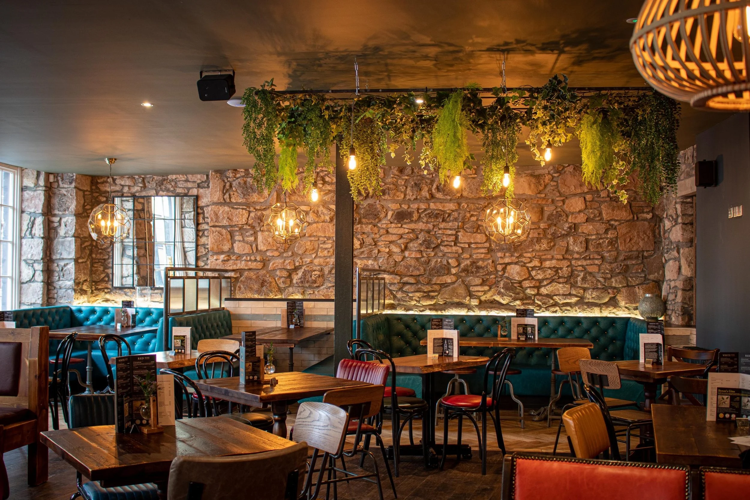 Interior of a cozy restaurant with exposed brick walls, green tufted banquettes, wooden tables, black chairs, hanging pendant lights, and green plant decor hanging from the ceiling.