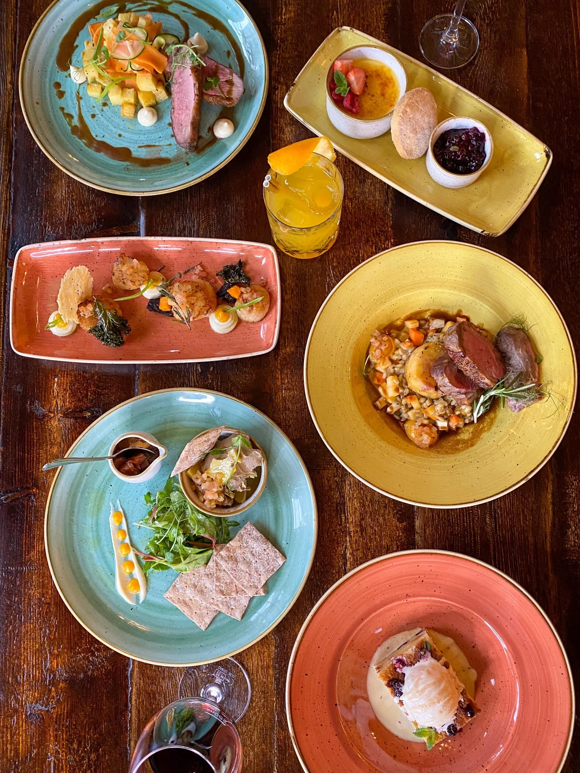 Assorted dishes from a fine dining meal, including salads, a main course with meat and vegetables, desserts, and drinks on colored plates on a wooden table.