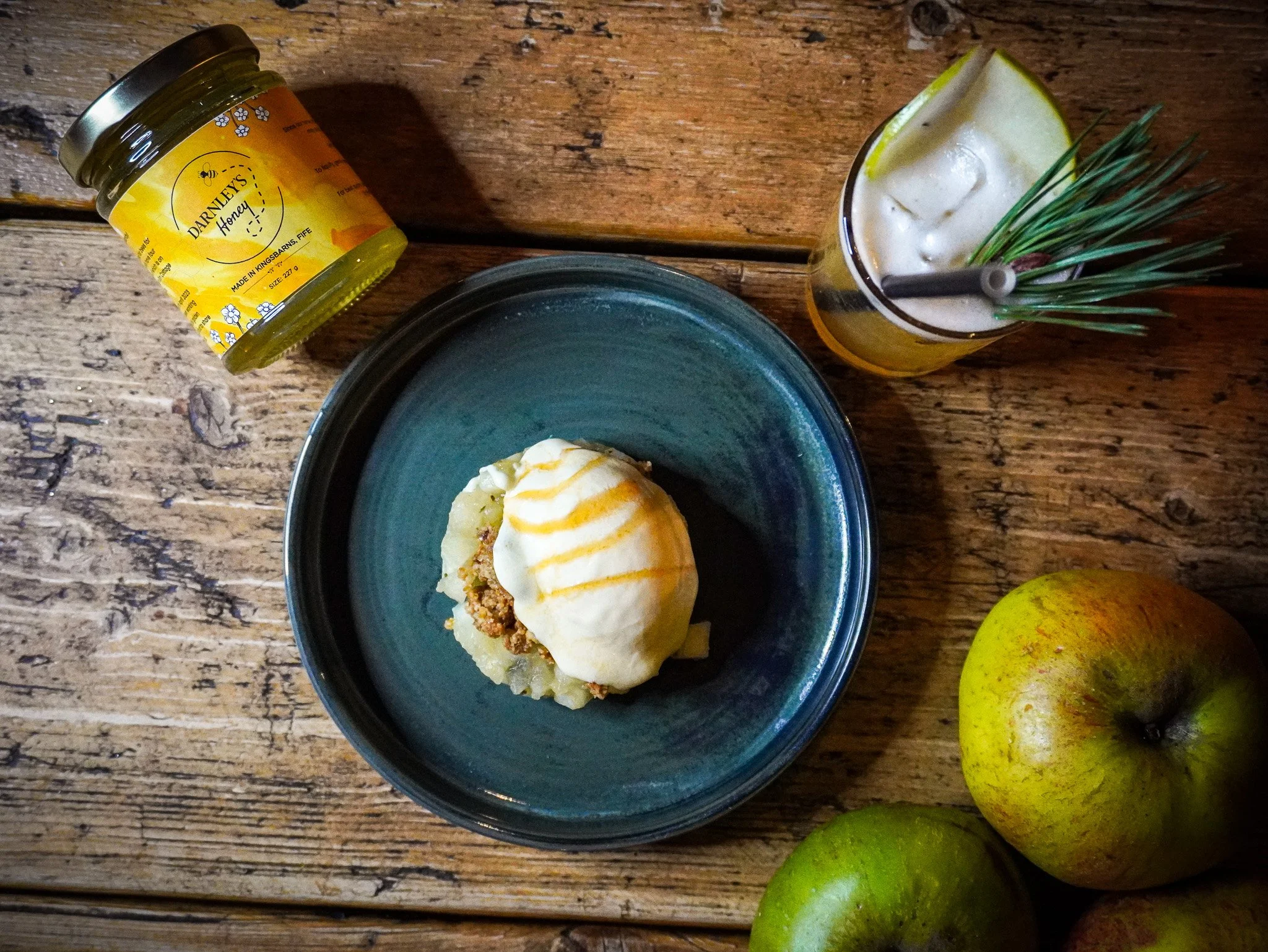A top-down view of a rustic wooden table with a bowl of shepherd's pie topped with mashed potatoes and gravy, a glass of lemonade garnished with a lemon wedge and pine needles, a jar of honey, and two green apples.