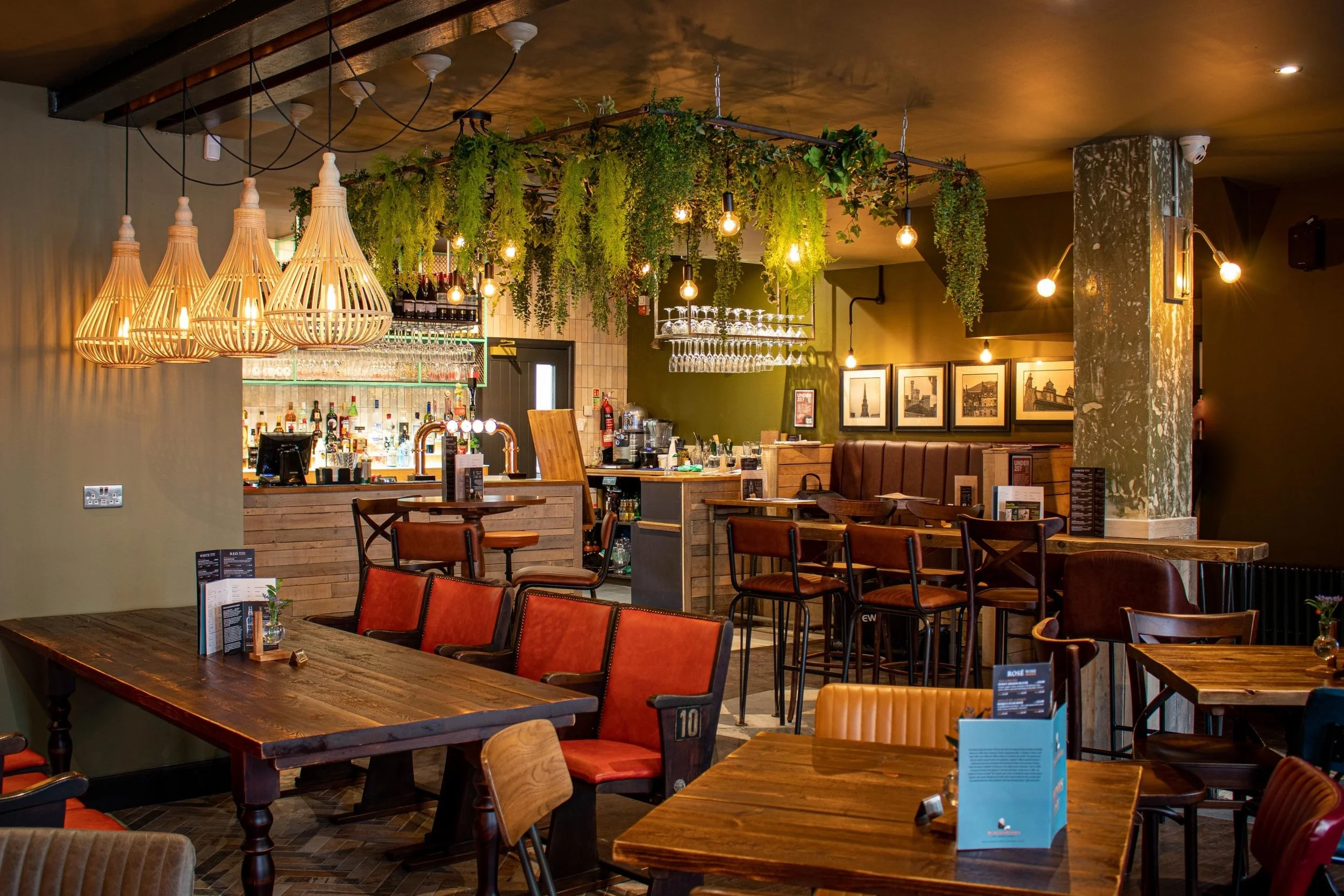 Interior of a cozy restaurant or bar with wooden tables and chairs, hanging wicker lamps, and greenery hanging from the ceiling, with a well-stocked bar in the background.