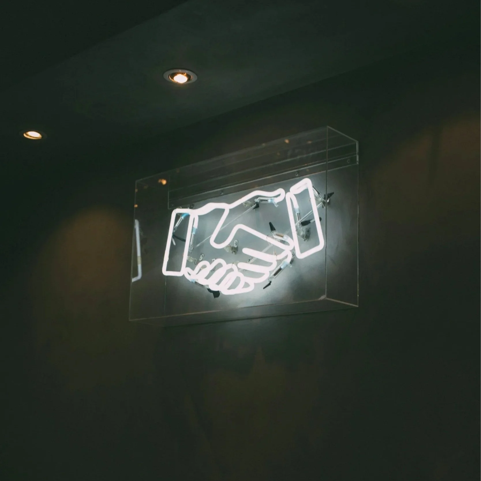 Neon sign of a handshake inside a clear rectangular case on a dark wall.