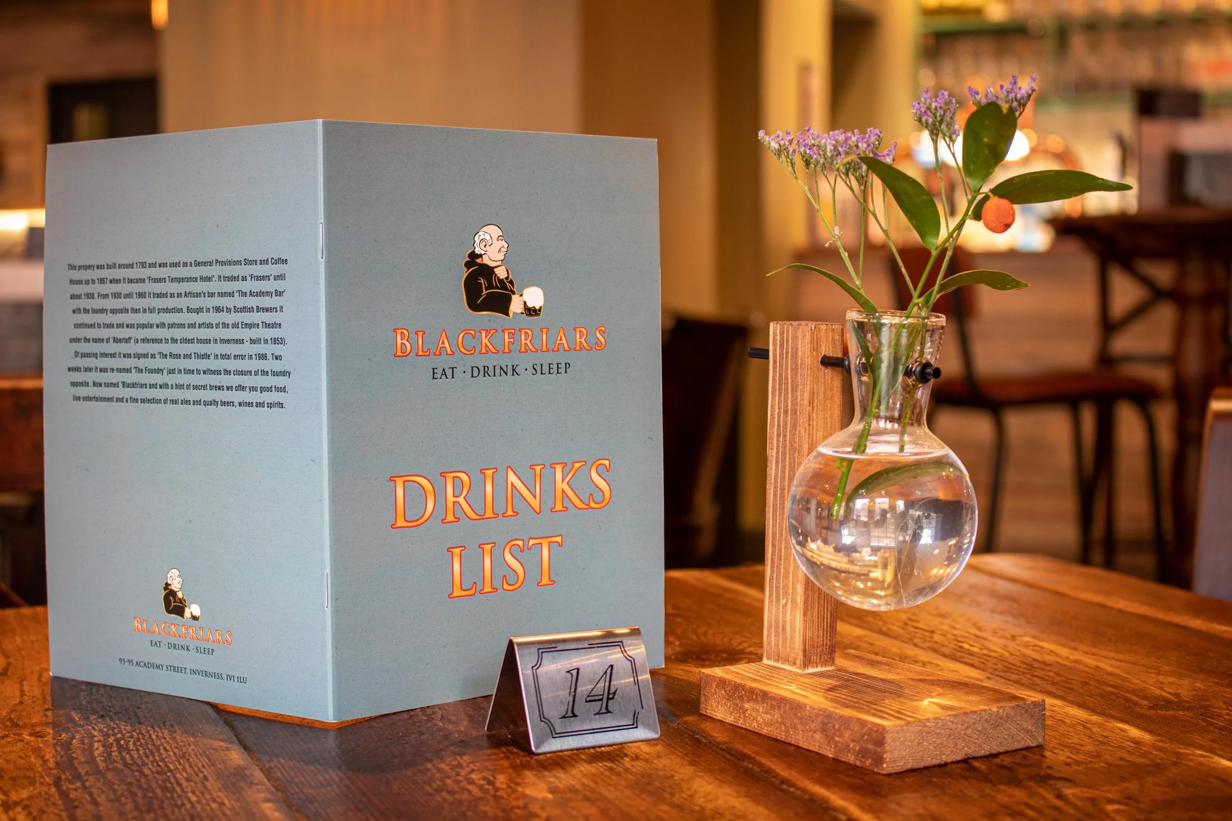 A drink menu booklet and a small table number sign are on a wooden table in a restaurant. The booklet is blue with orange and black text, with the logo of a bearded man holding a mug, and the words "BLACKFRIARS EAT DRINK SLEEP" and "DRINKS LIST" on t