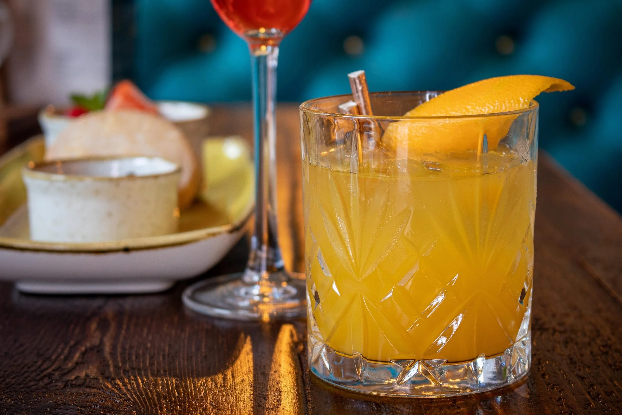 Close-up of a glass of yellow cocktail with lemon wedge and cinnamon stick, with a plate of dip and snacks in the background.