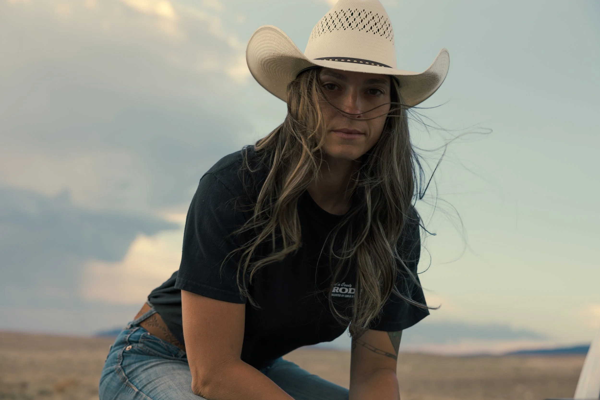 A woman wearing a large white cowboy hat, a black T-shirt, and blue jeans is outdoors in a rural or open landscape with a cloudy sky in the background.
