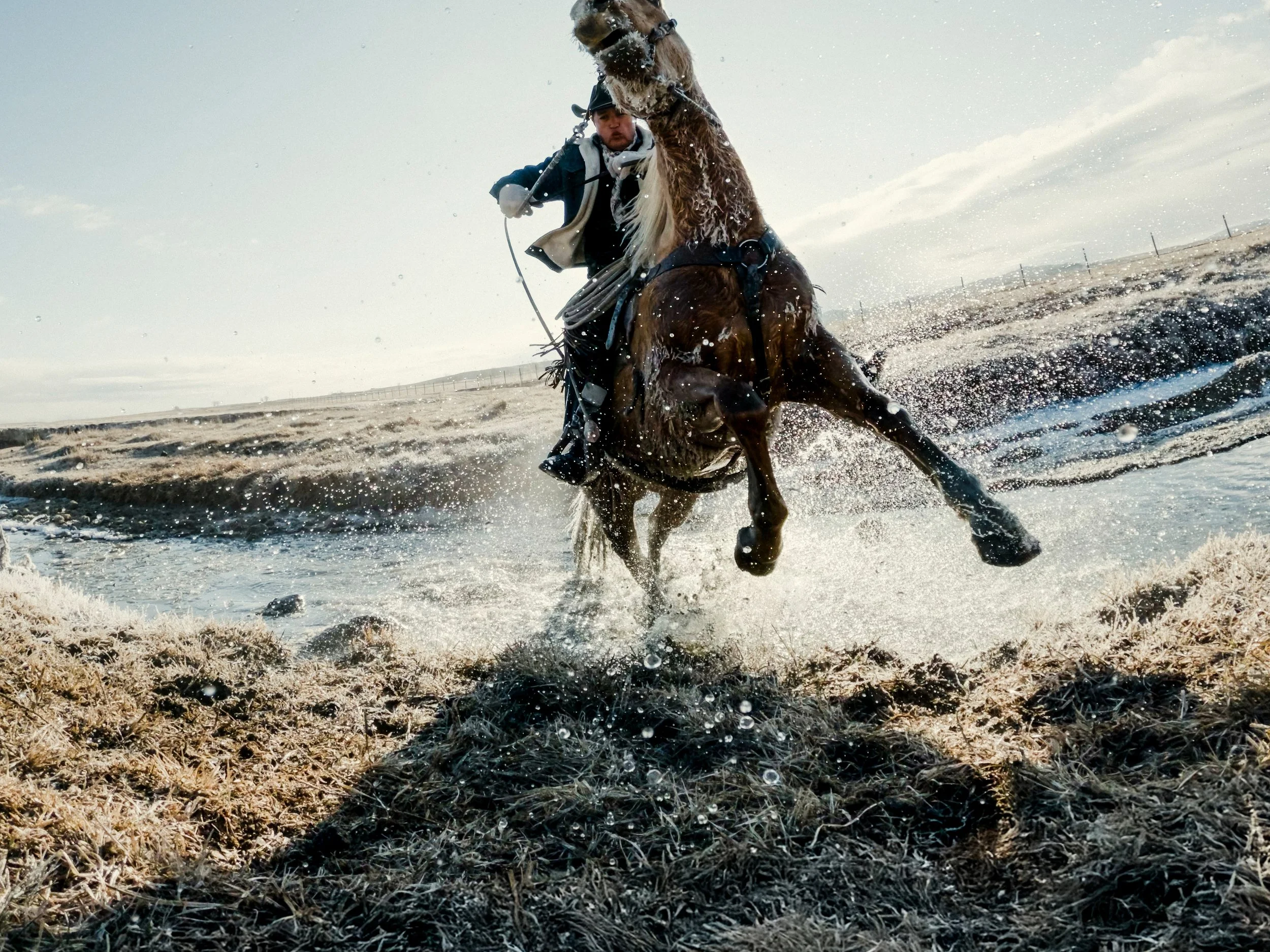 A man riding a boat on a horse through a water crossing on a rugged, grassy terrain under a cloudy sky.