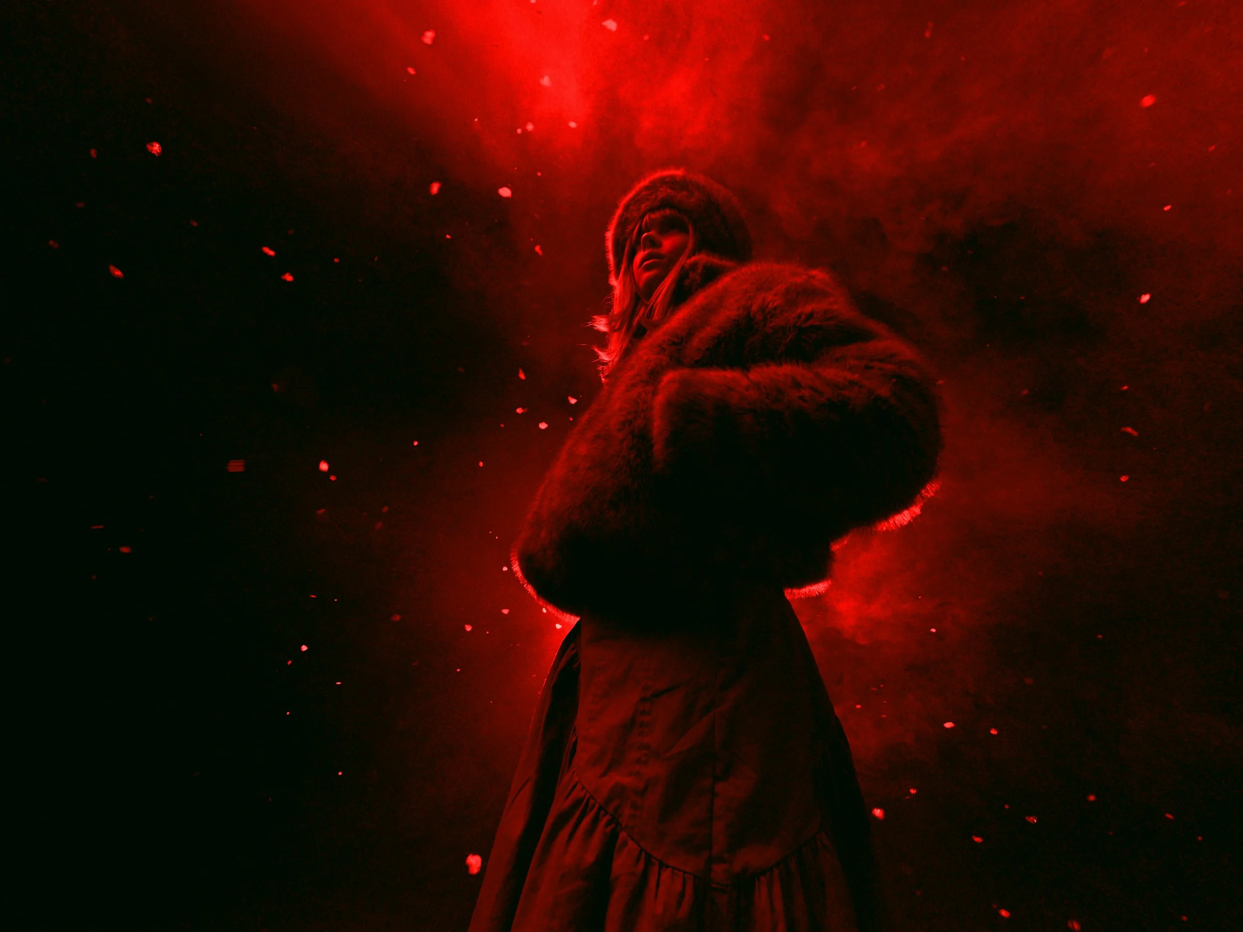 A person dressed in a fur coat and wearing a hat, standing with arms crossed against a dramatic red and black background with floating particles.