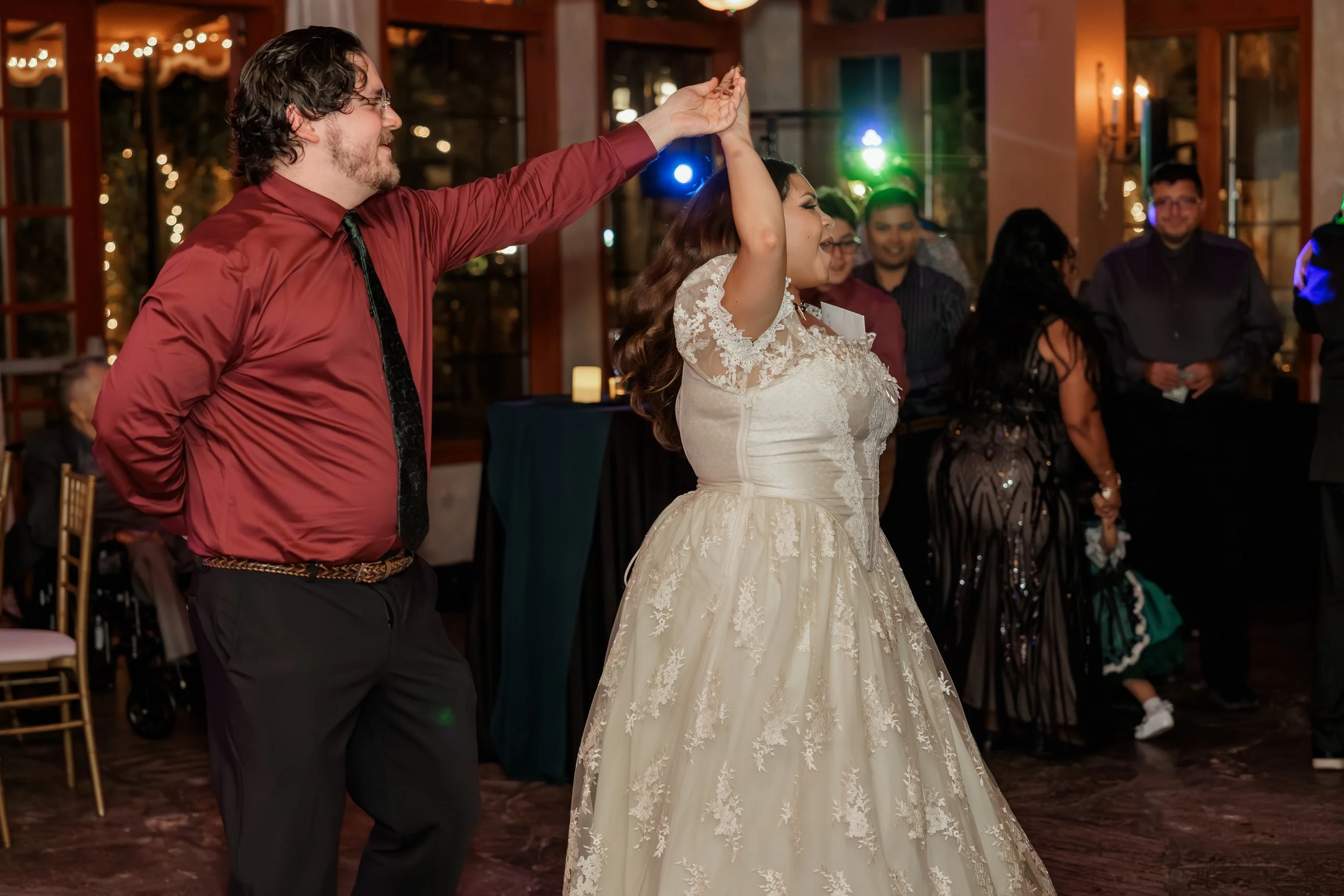A man with curly hair and glasses wearing a red shirt and black tie dances with a woman in a white lace wedding dress. They are in a warmly lit venue with other people in the background, some smiling and watching, during a celebration or wedding rece