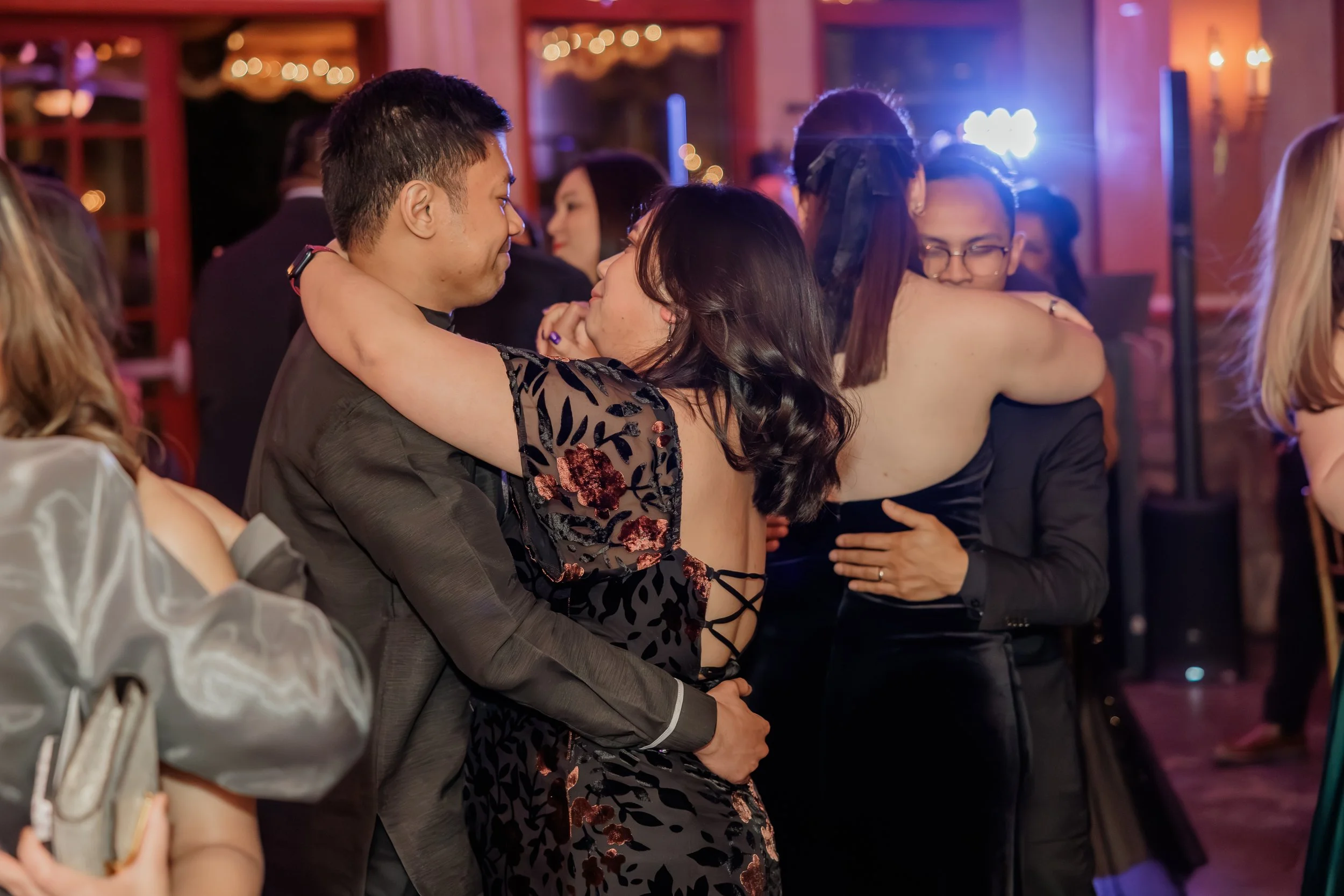 Couple dancing closely at a party, with other guests around them, in a warmly lit room with festive decorations.