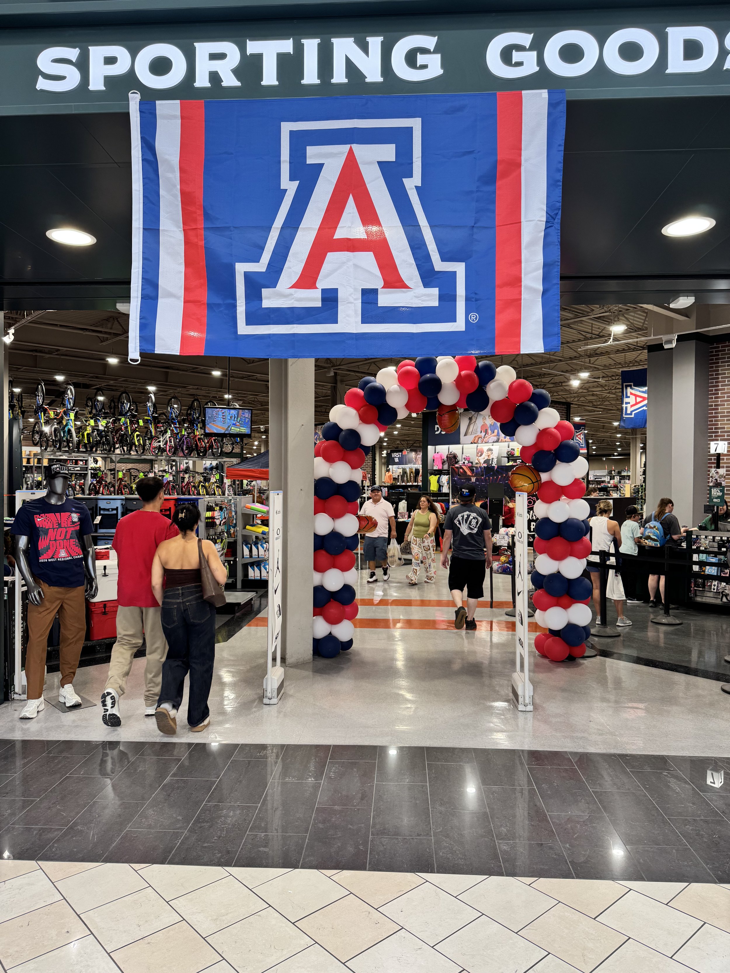 Entrance to an athletic store decorated with red, white, and blue balloons, featuring a large University of Arizona flag and a group of shoppers inside.